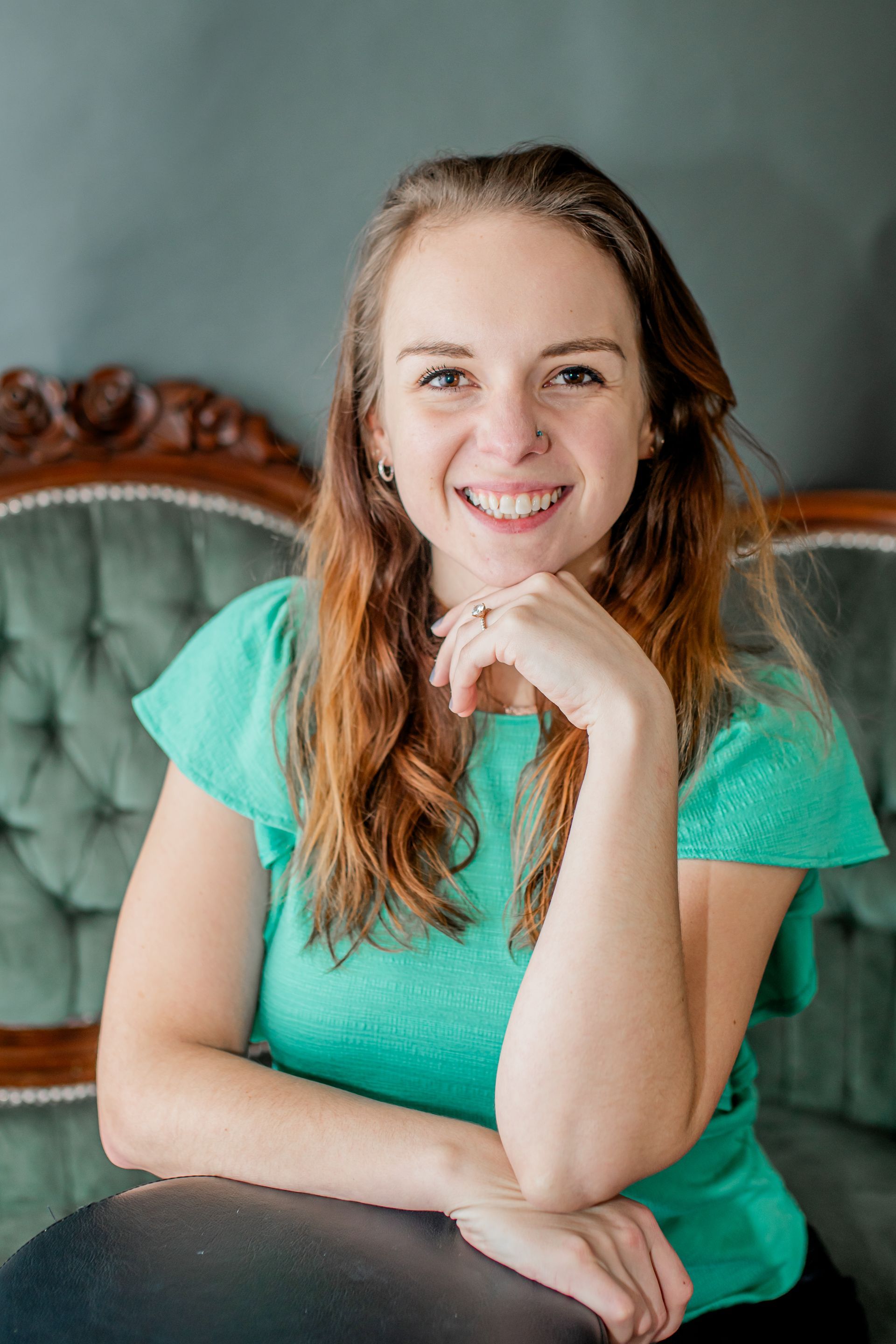 Smiling woman in a turquoise shirt seated on a vintage green chair, resting her chin on her hand.