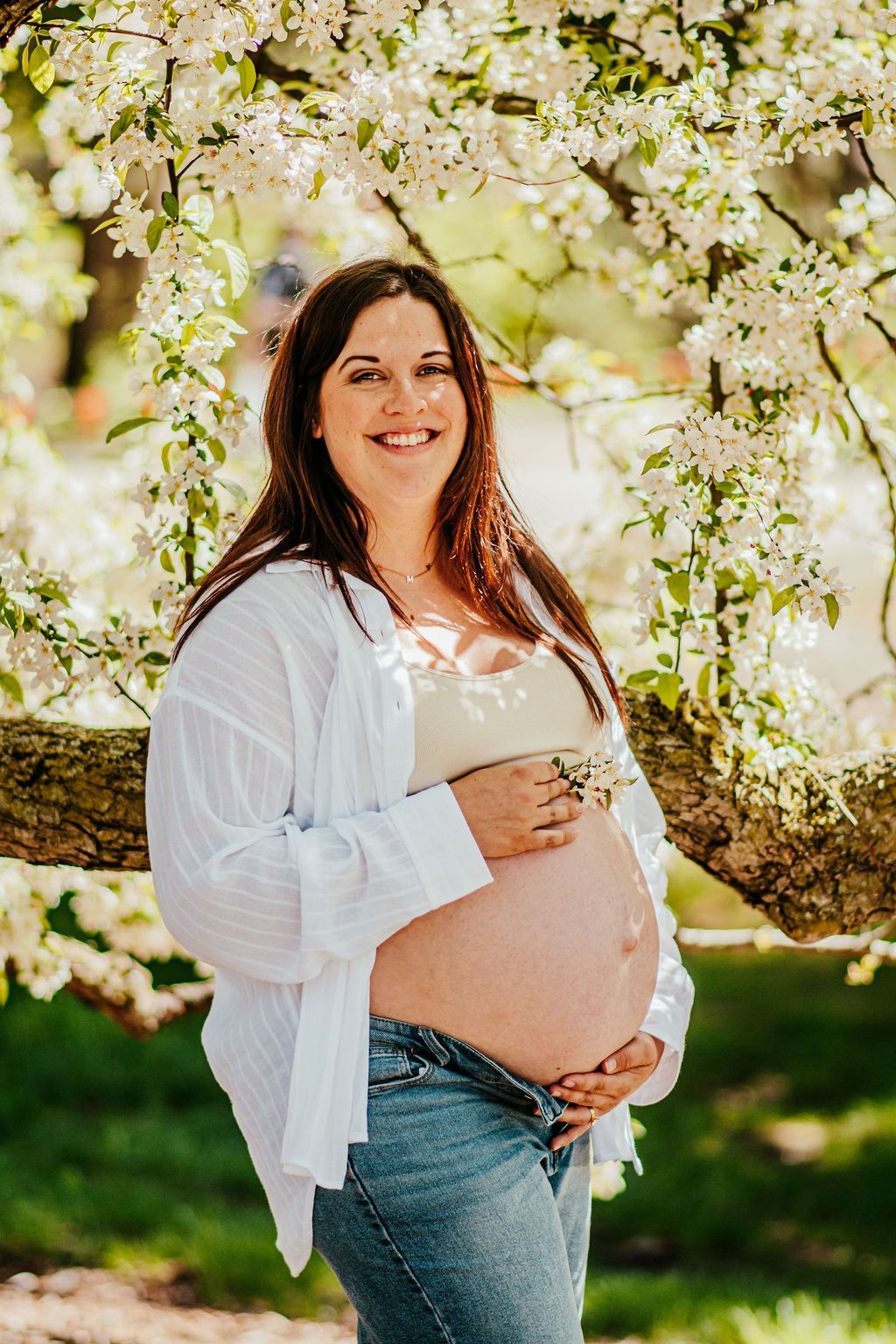 Pregnant woman smiling outdoors under blooming tree branches, holding her belly.