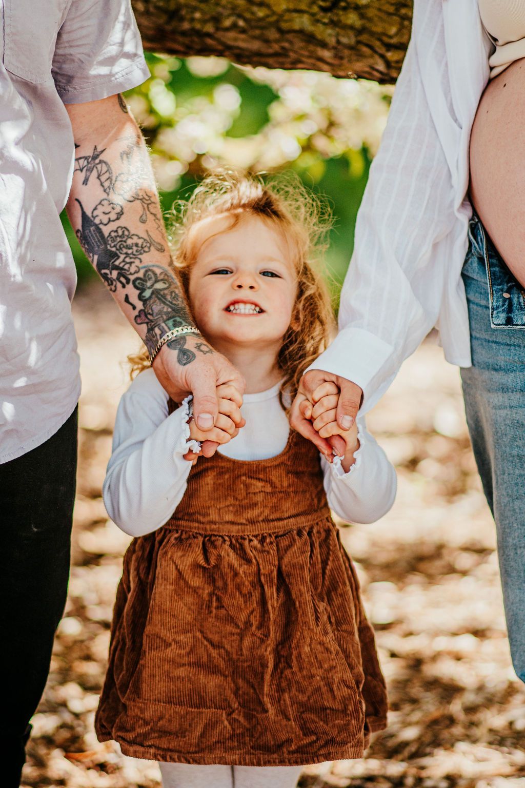 Child in brown dress holding hands with two adults outdoors in dappled sunlight