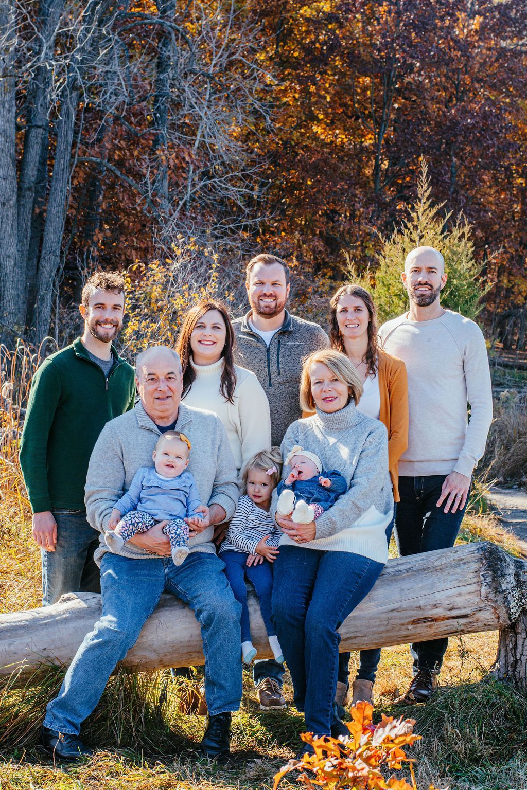 Family seated outdoors on a log, smiling in a sunlit autumn forest.