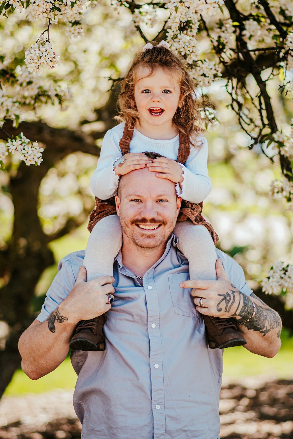 Smiling man carrying a child on his shoulders in a sunlit orchard, with blossoming trees behind them