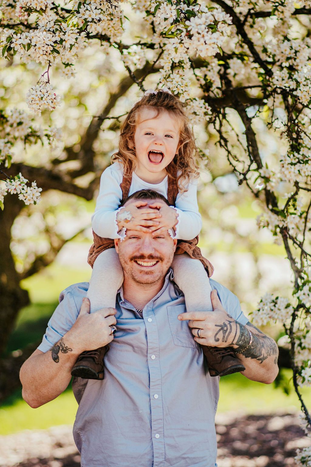 Man smiling while carrying a child on his shoulders under blooming tree branches
