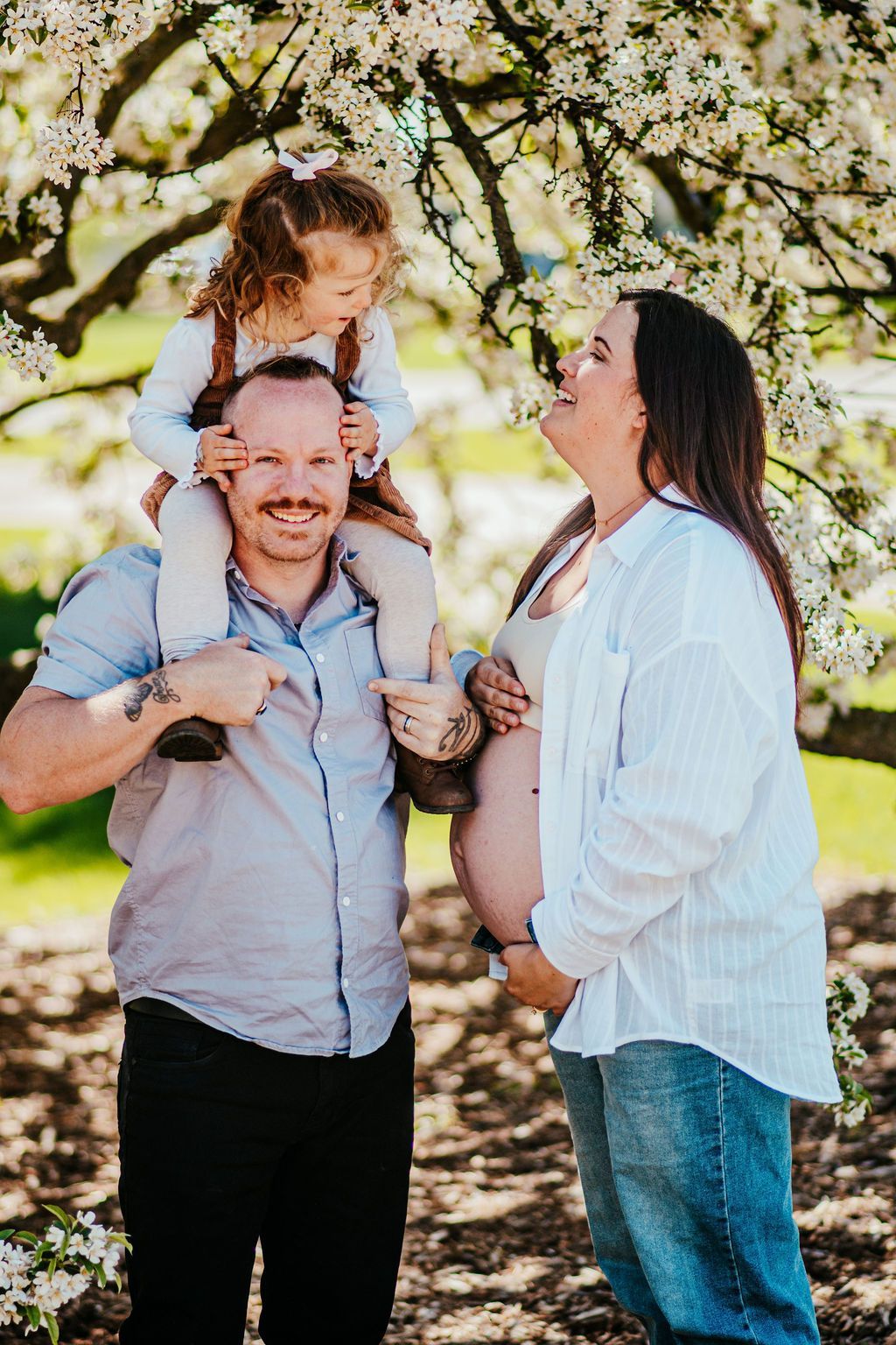 Family outdoors under blooming trees, smiling and embracing in soft sunlight.