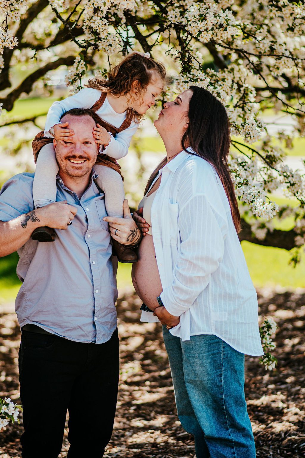 Family standing under blooming tree, smiling while a child sits on a man’s shoulders and another person kisses his cheek