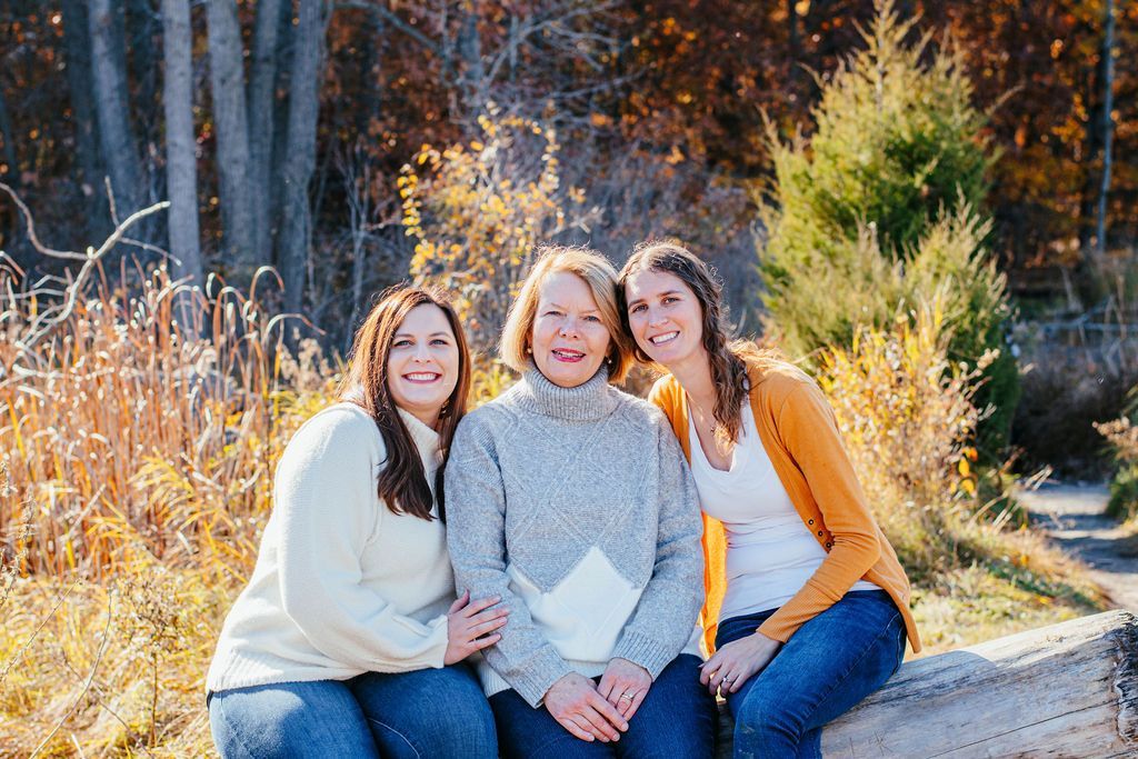 Three people sitting outdoors in autumn, smiling beside tall grasses and trees.