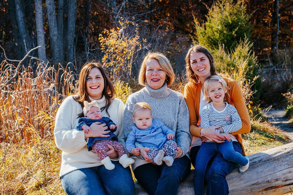 Four people sitting outdoors in a sunlit garden, smiling and holding two children.