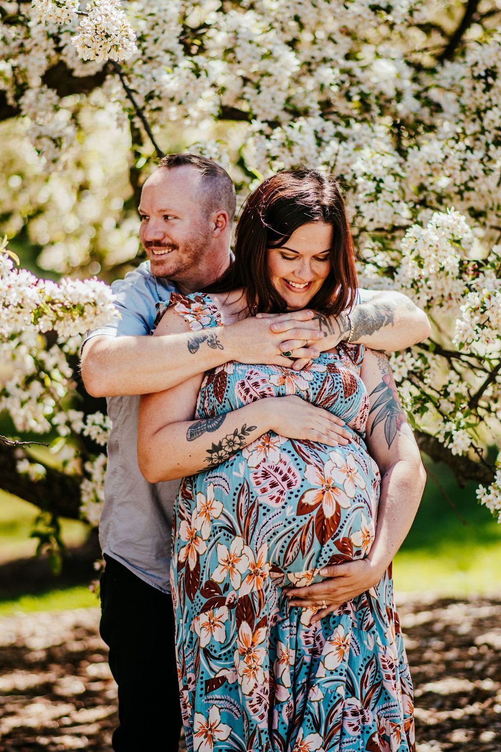 Couple embracing outdoors under blooming white blossoms, smiling warmly, with one partner visibly pregnant