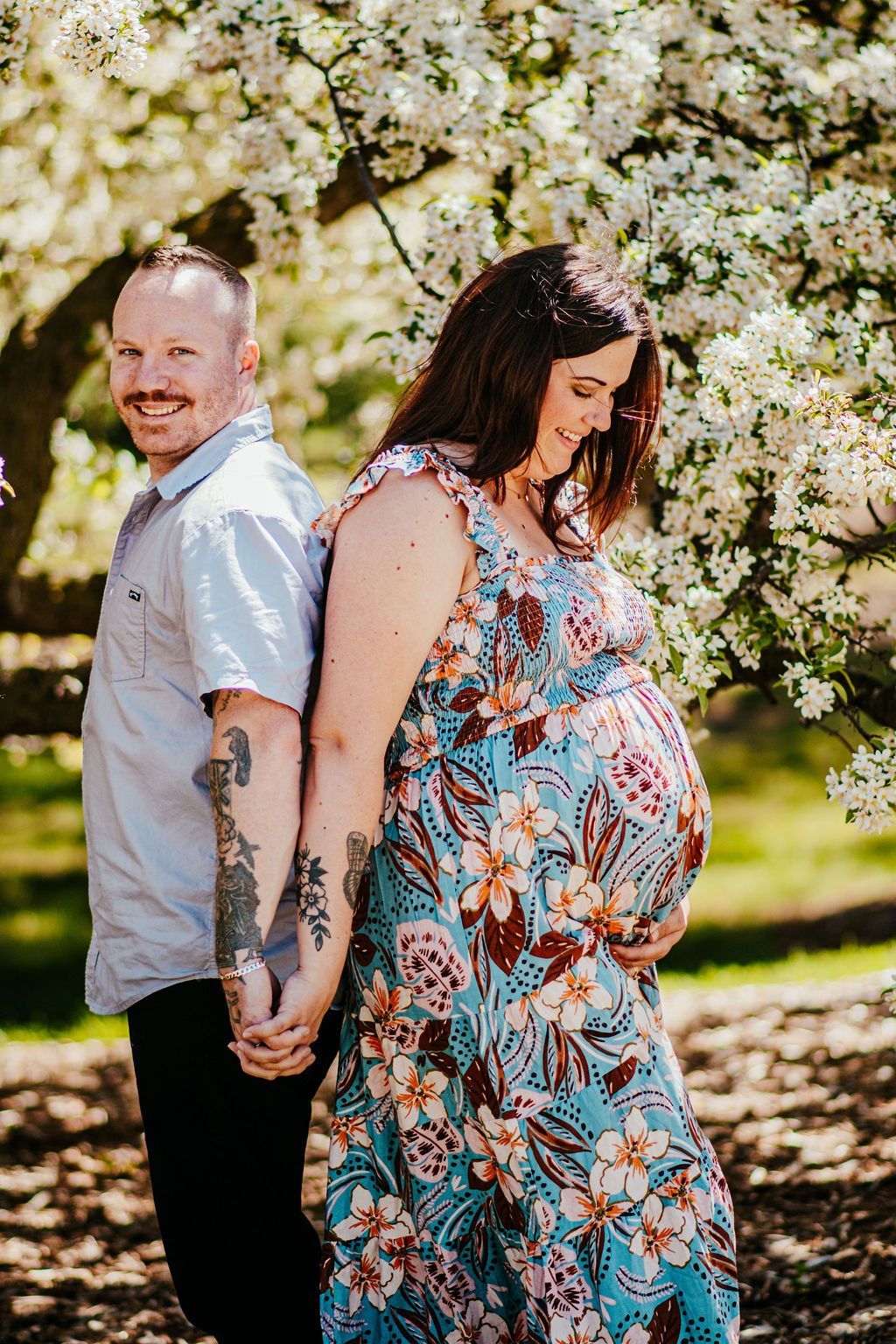 Couple standing back-to-back and holding hands in a blooming garden, with the woman in a floral dress.
