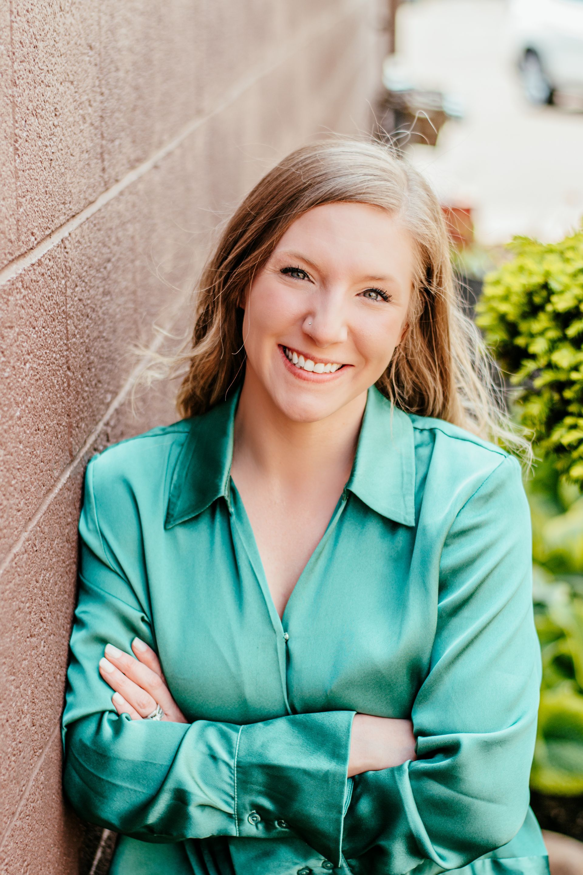 Smiling person in a teal shirt leaning against a wall outdoors with arms crossed