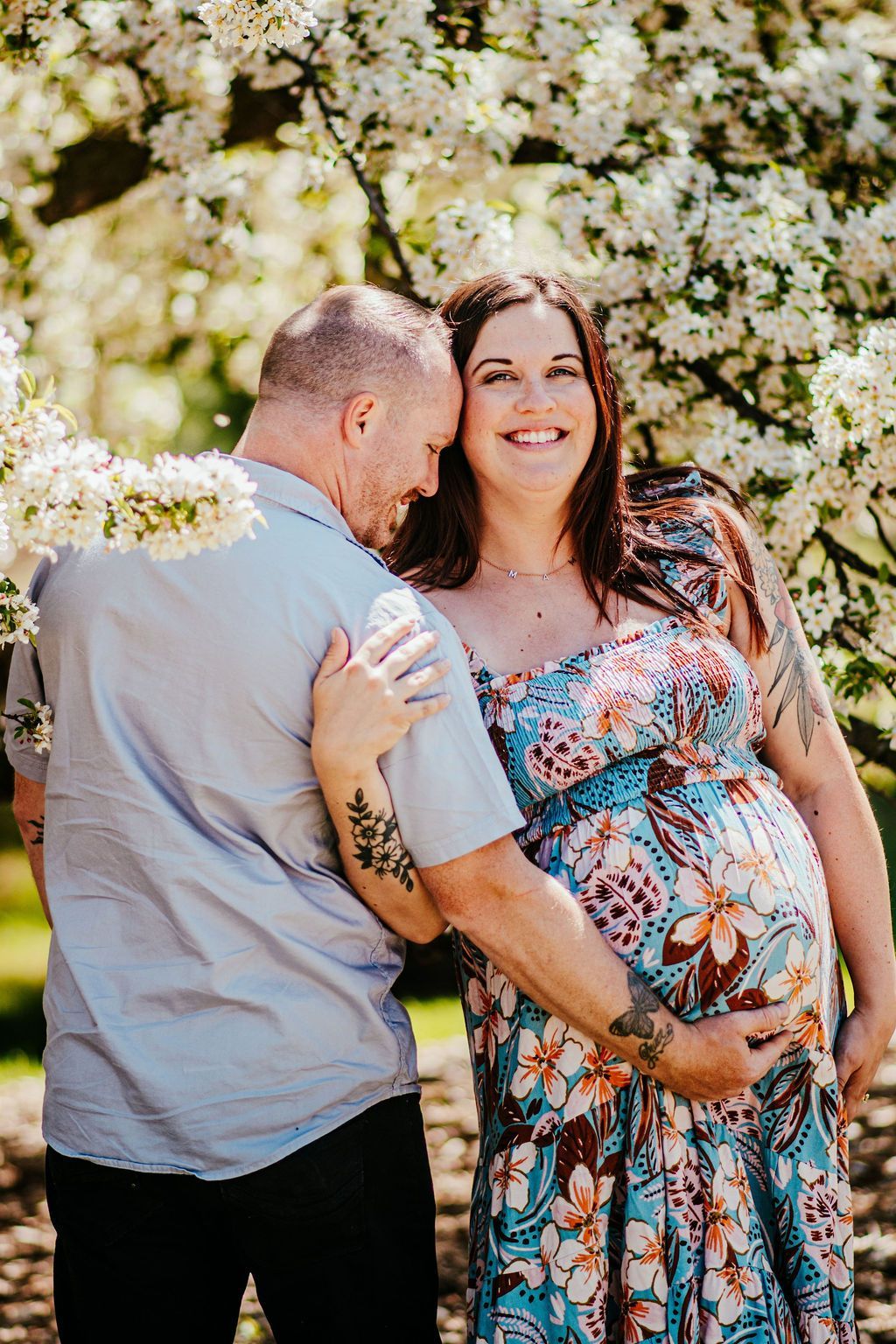Couple standing under white blossoms, smiling and embracing, with a pregnant woman in a blue floral dress