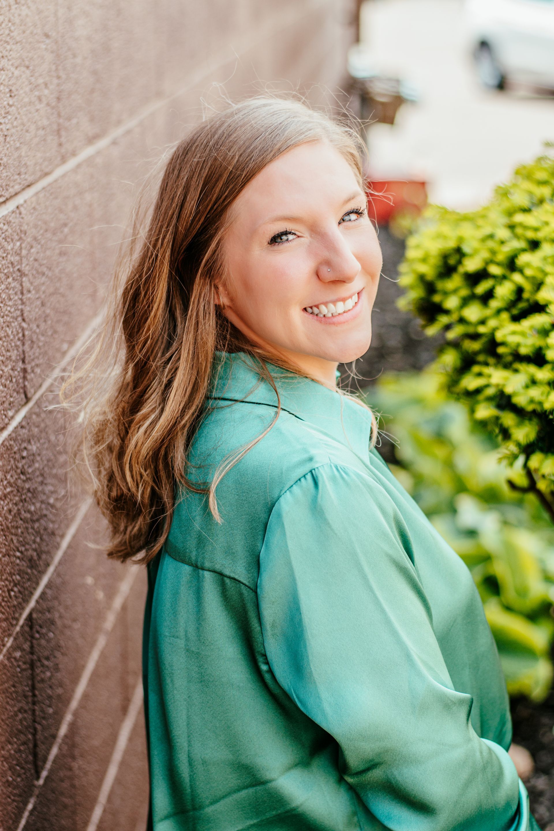 Woman in a green blouse smiling over her shoulder beside a brick wall and greenery