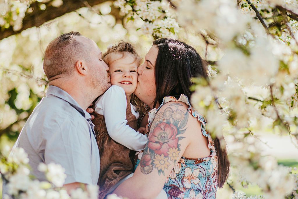 Three people embracing outdoors under sunlit trees, one adult kissing a child’s cheek.