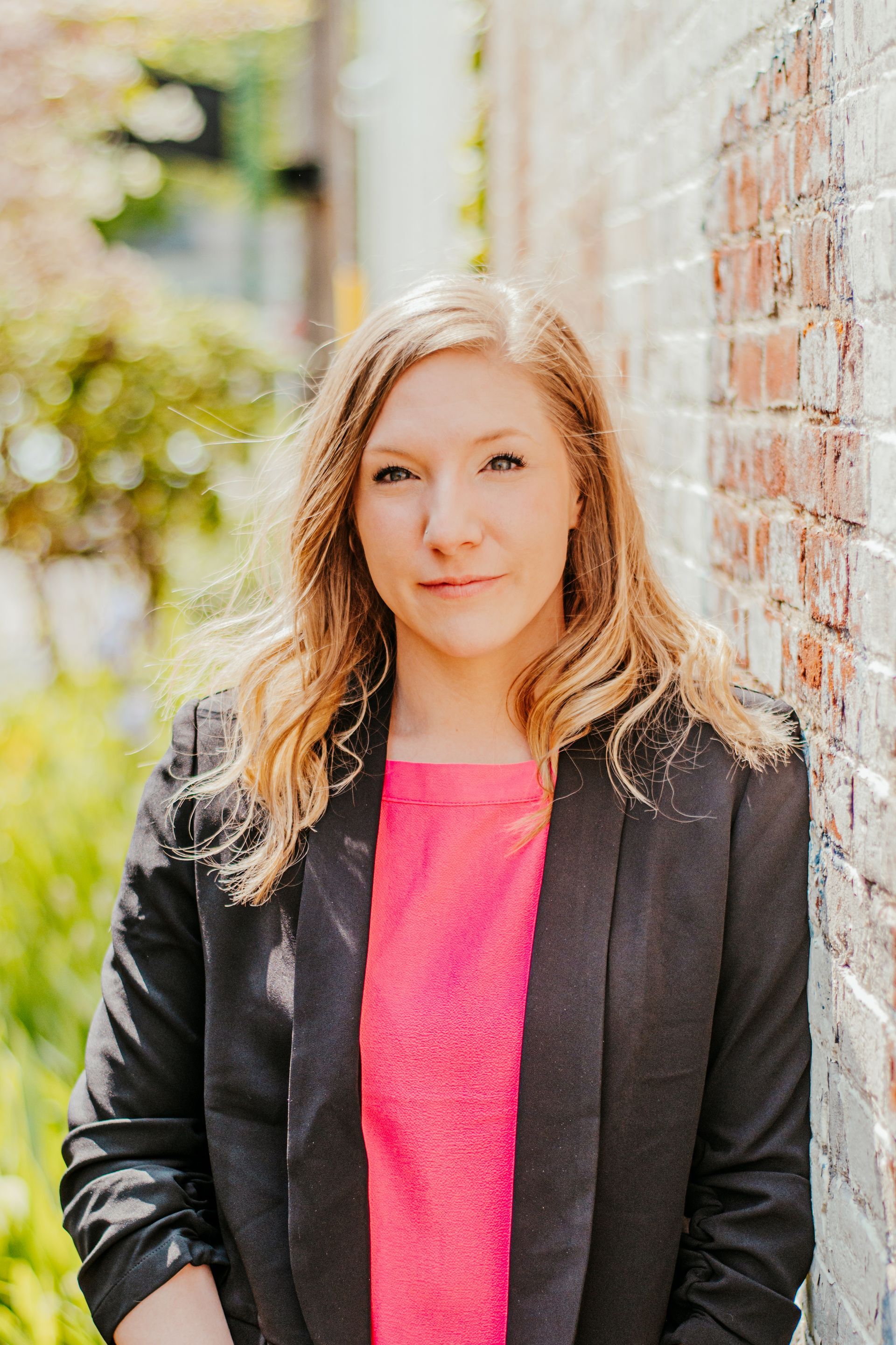 Woman in a black blazer and bright pink top standing beside a brick wall outdoors