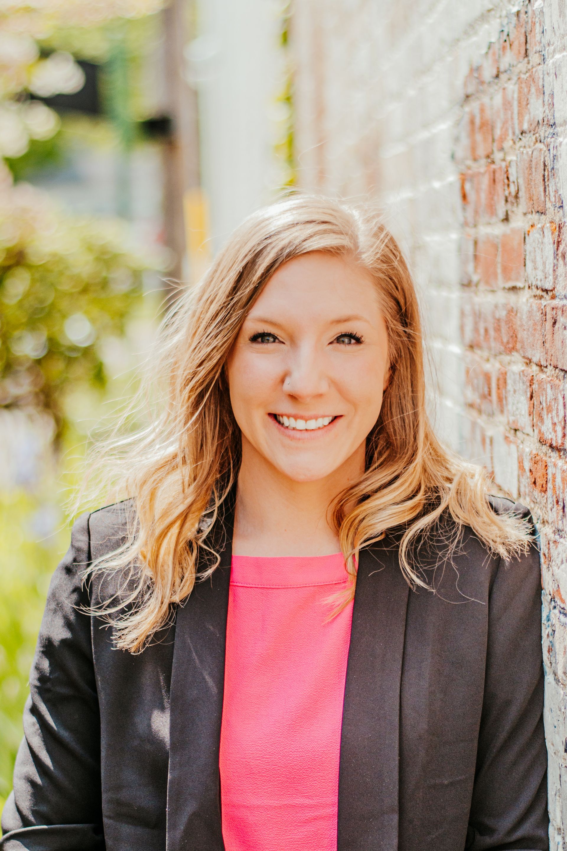Smiling woman in a dark blazer and pink top stands beside a brick wall outdoors.