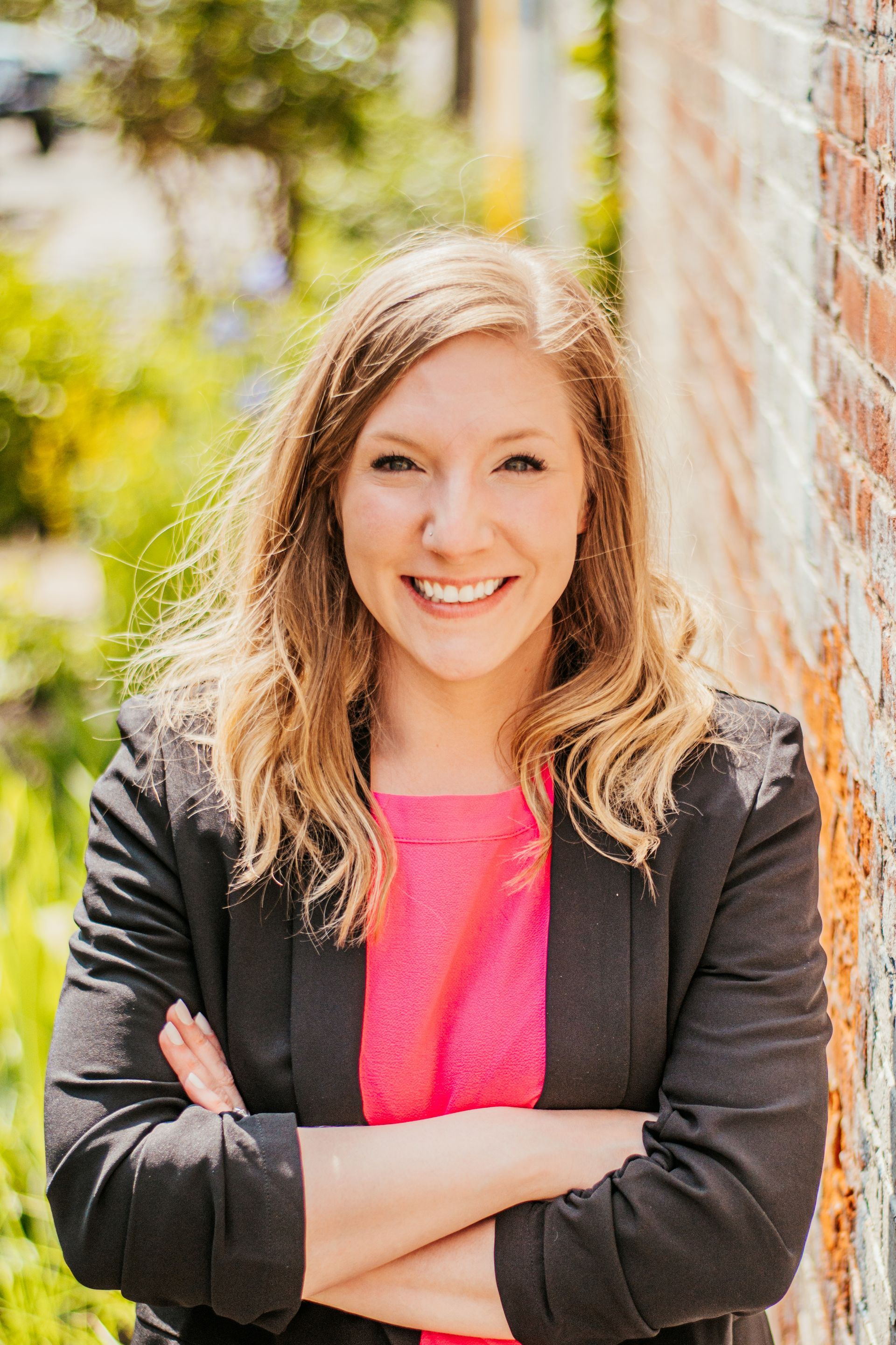 Smiling woman with crossed arms in a black jacket and pink shirt standing outdoors by a brick wall