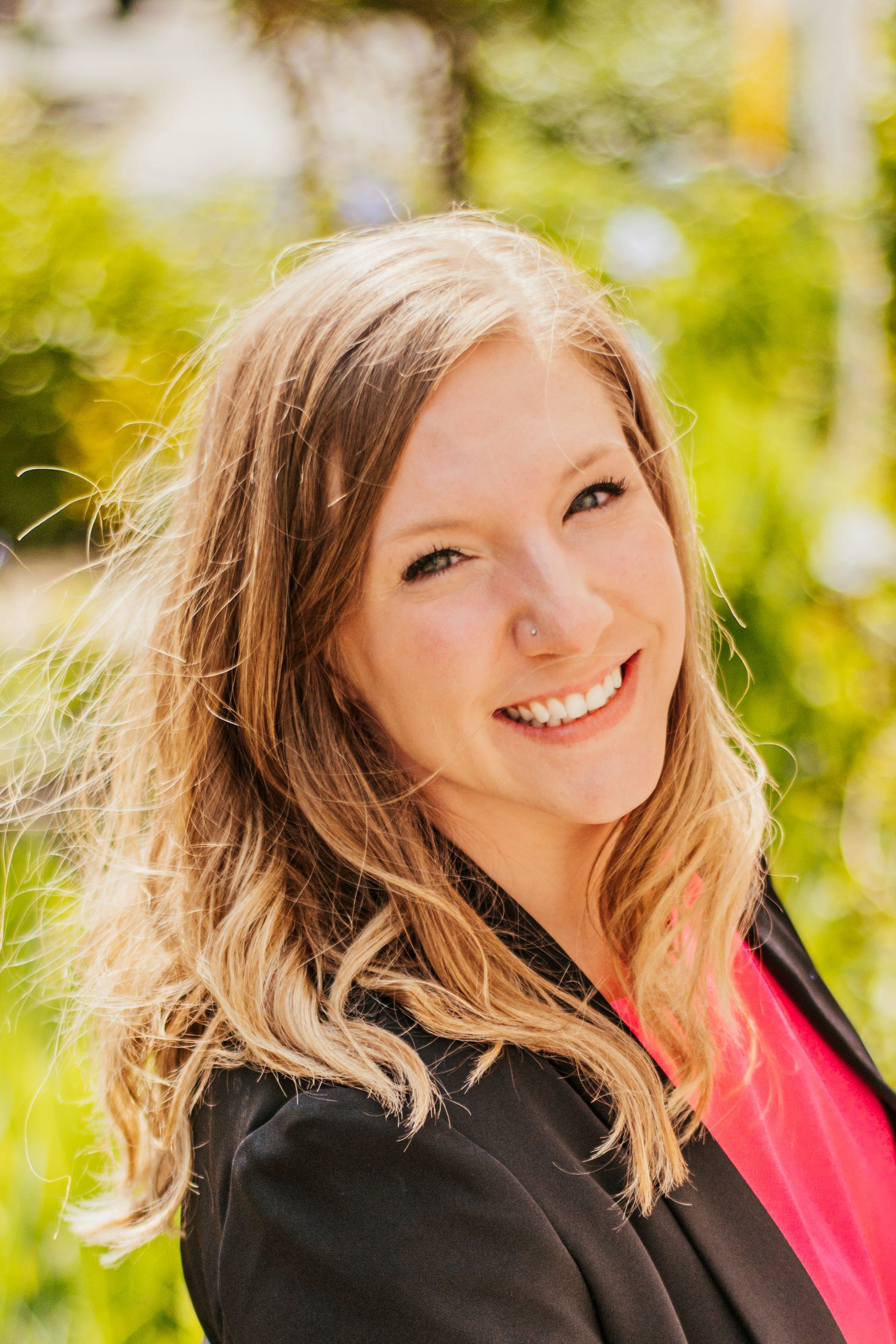 Smiling woman with long blonde hair outdoors, wearing a black jacket and pink top.