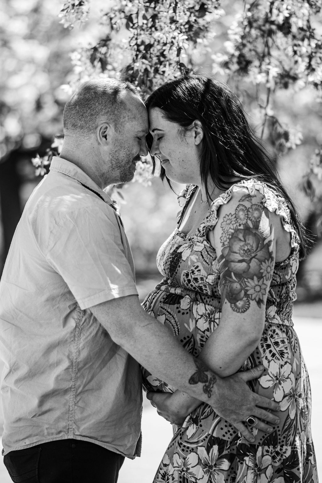 Couple touching foreheads and holding hands in black-and-white, standing outdoors with blooming trees behind them