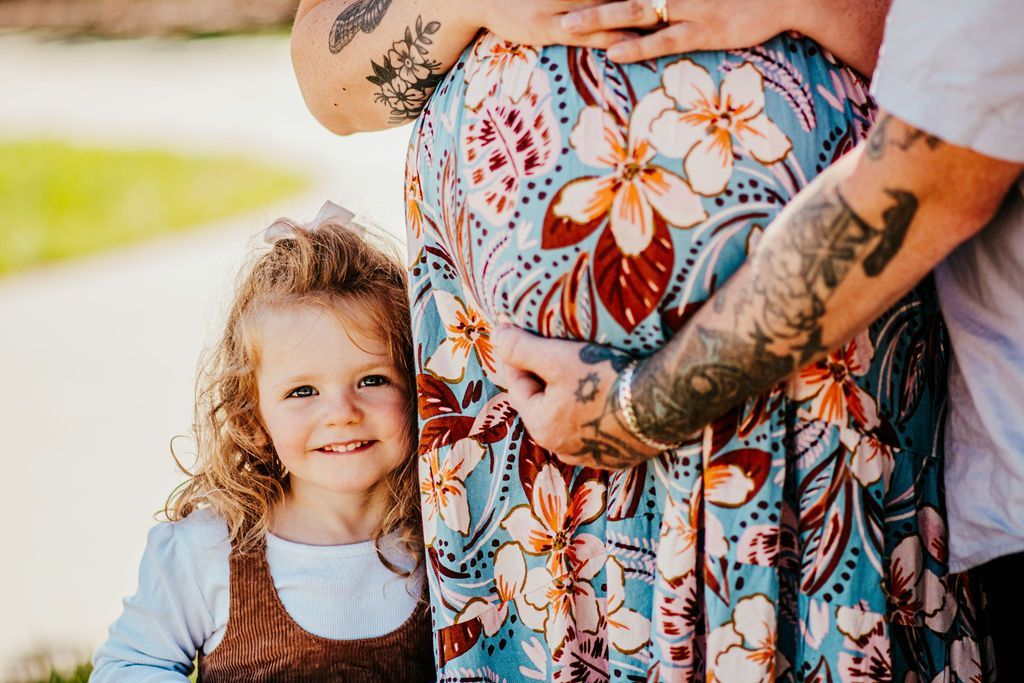 Pregnant person in floral dress with tattooed arms, holding a young child outdoors