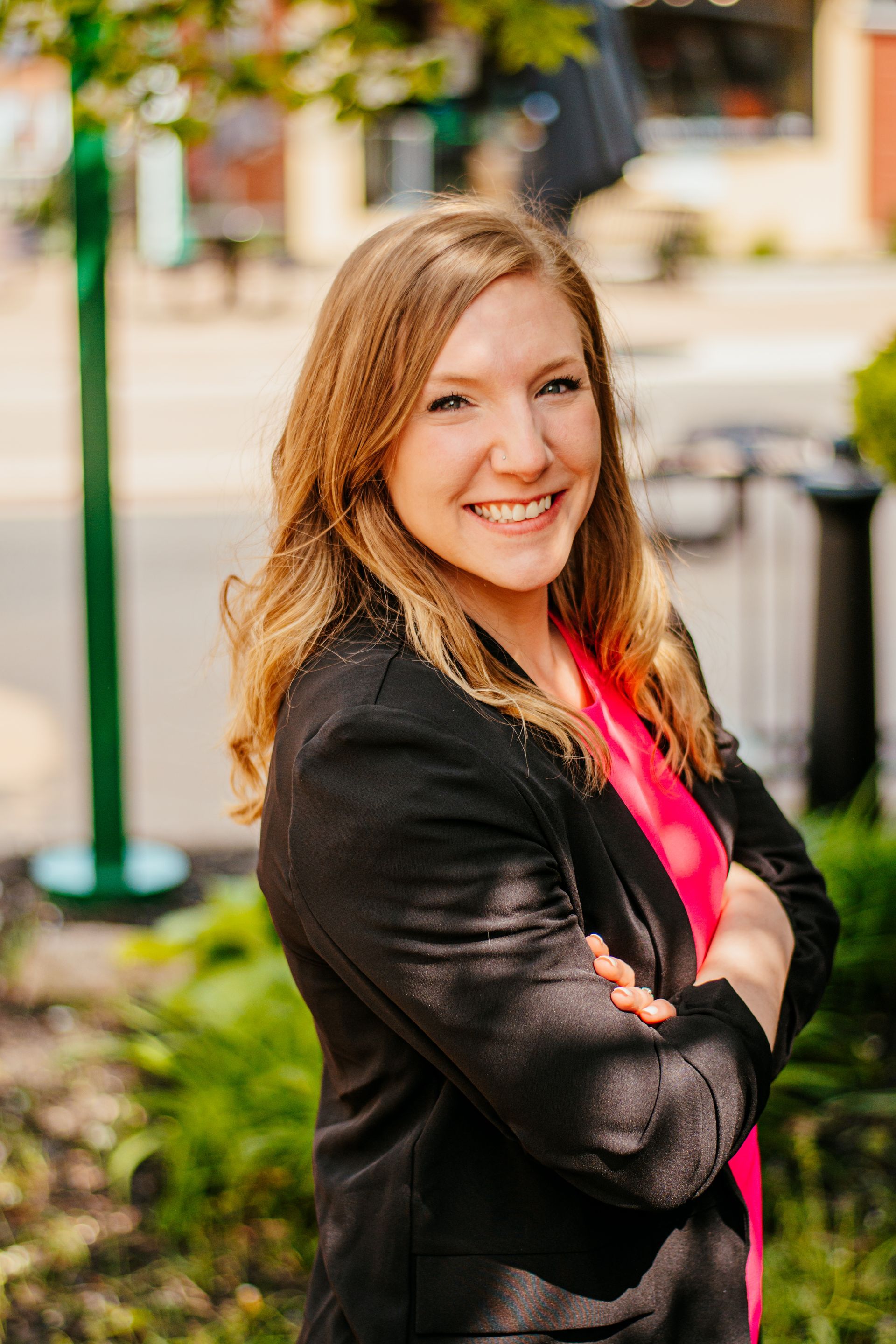 Smiling woman in a black jacket and pink top standing outdoors with arms crossed