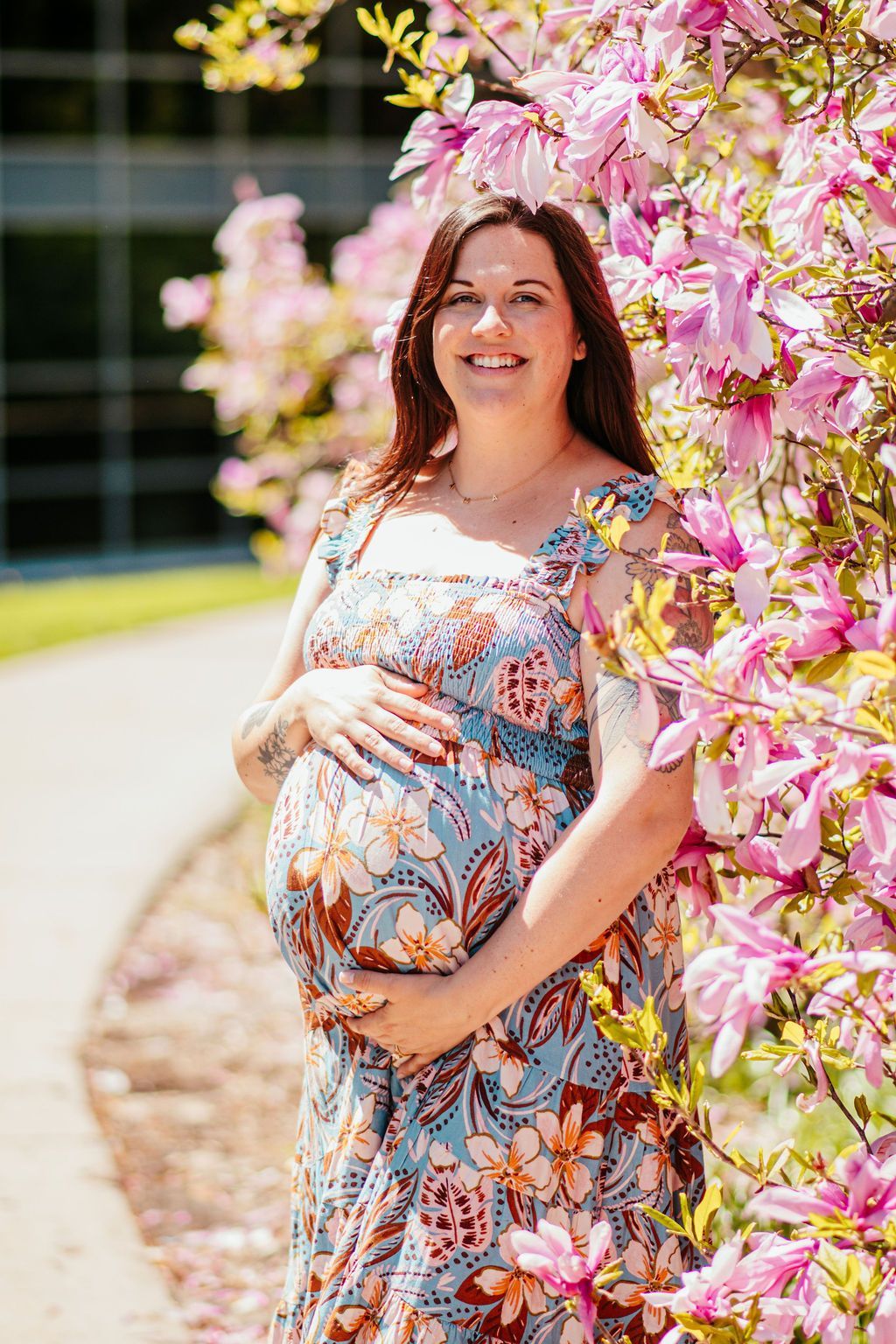 Pregnant person smiling beside blooming pink flowers outdoors, wearing a floral dress