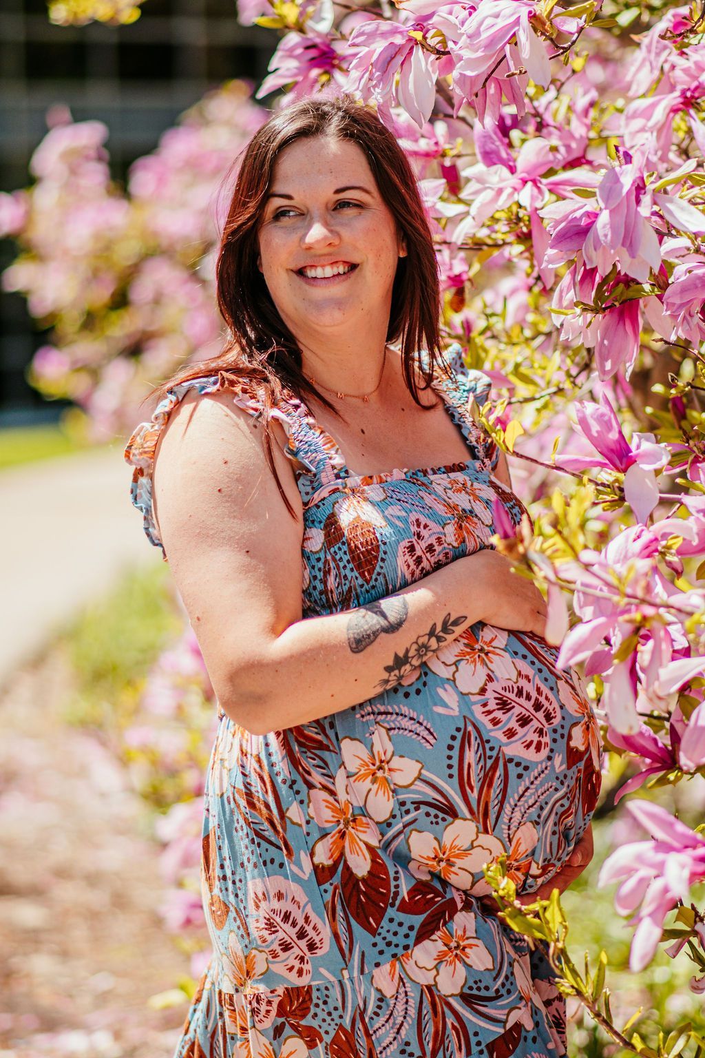 Woman in a floral dress standing by pink blossoms, smiling outdoors.
