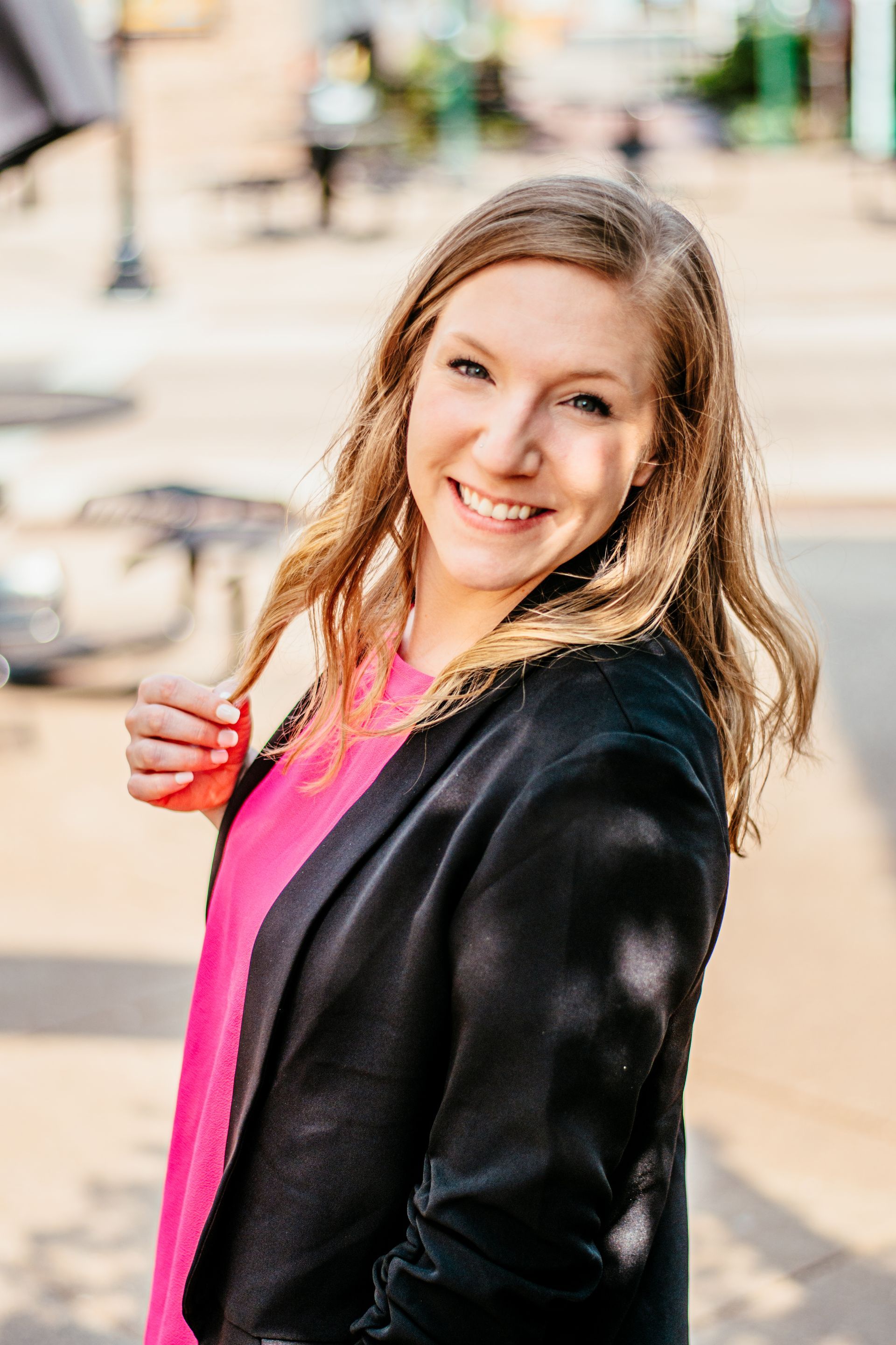 Smiling woman in a black jacket and pink top, standing outdoors on a city street, holding a bag strap.