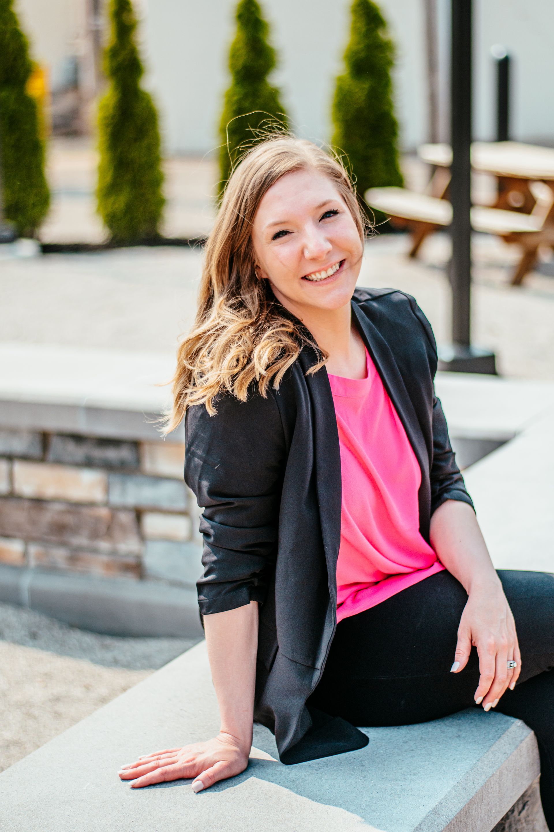 Smiling woman in a black blazer and pink top sitting on a stone ledge outdoors
