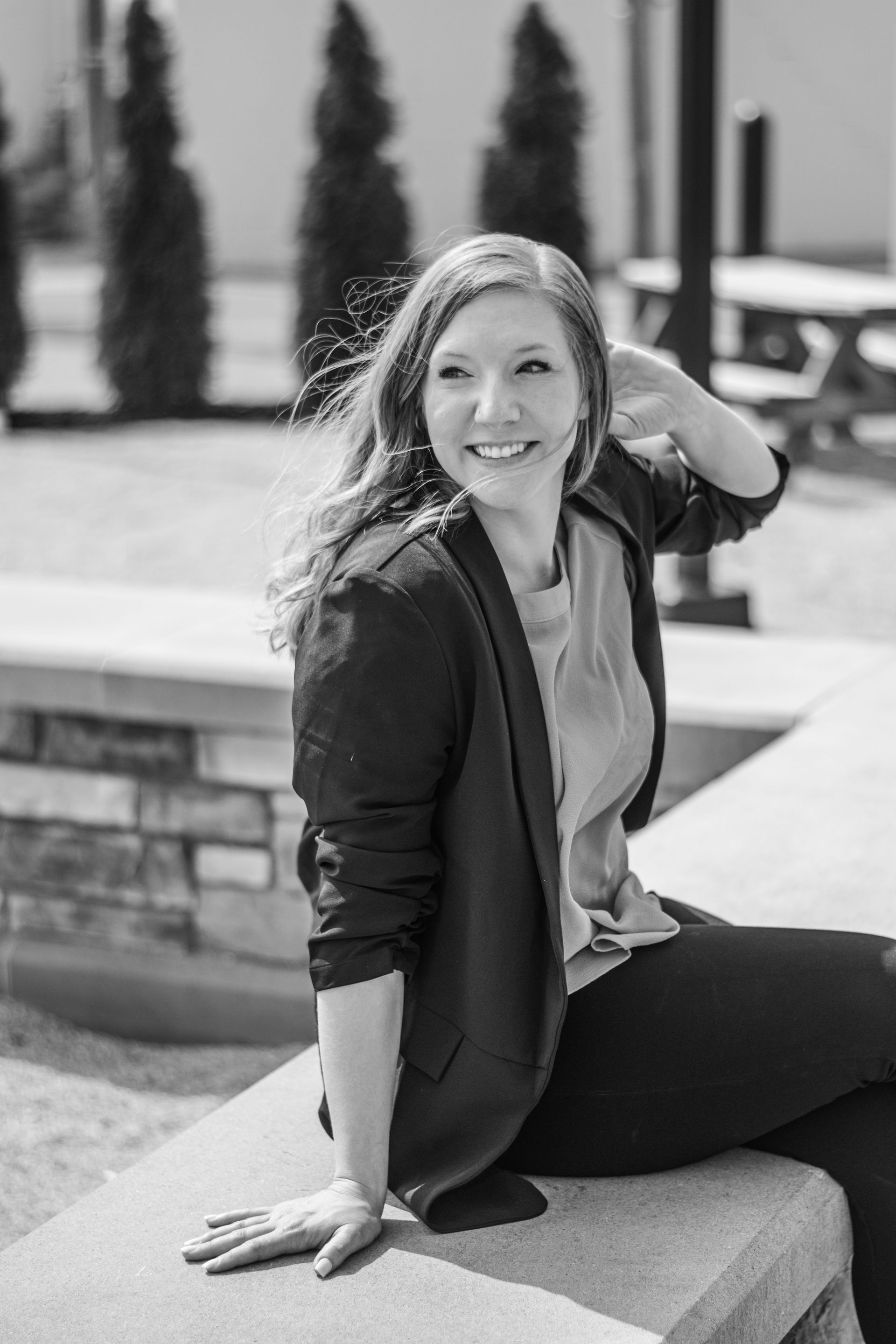 Woman sitting on a stone ledge outdoors, smiling with one hand behind her head.