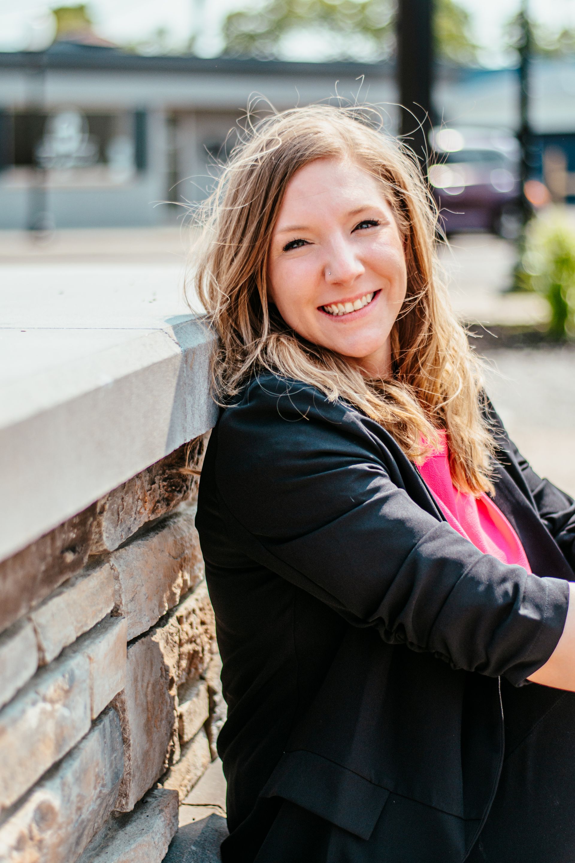Smiling woman in a black jacket leaning on a stone wall outdoors, with a blurred street background