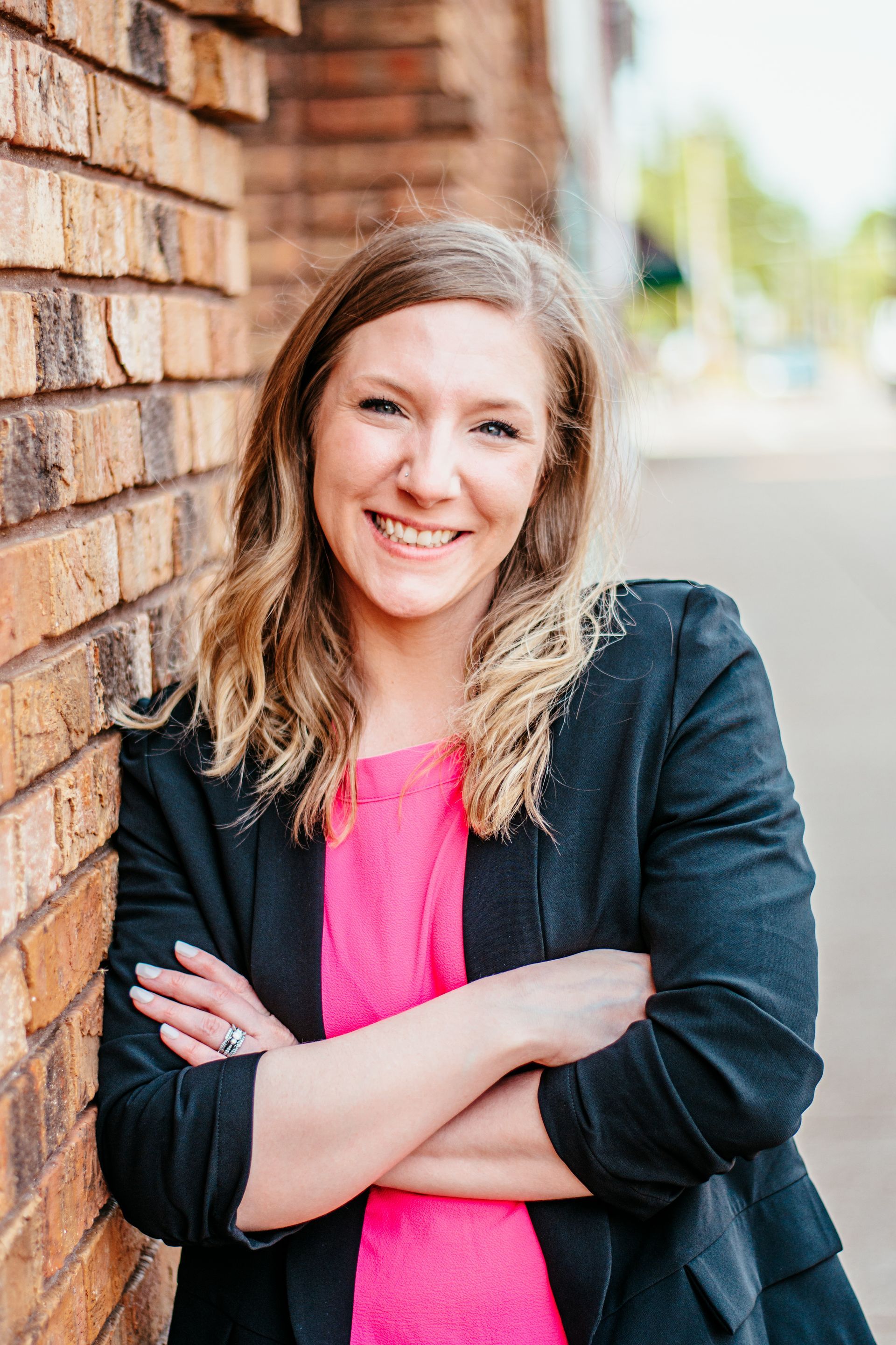 Smiling woman in a black jacket and pink top leaning against a brick wall outdoors