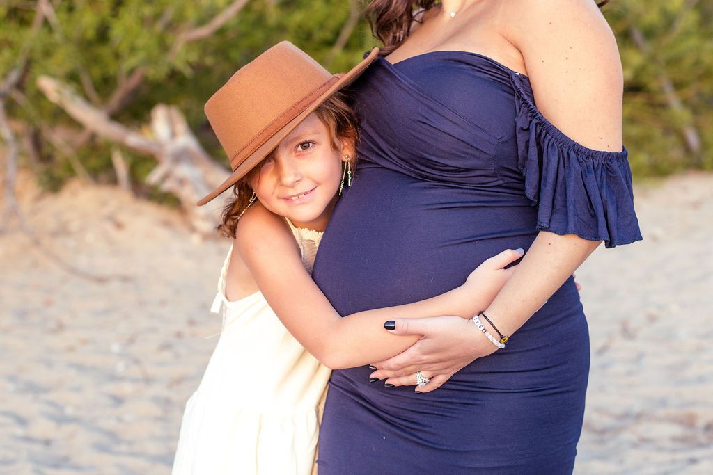 Pregnant woman in a navy dress hugging a child in a hat on a beach