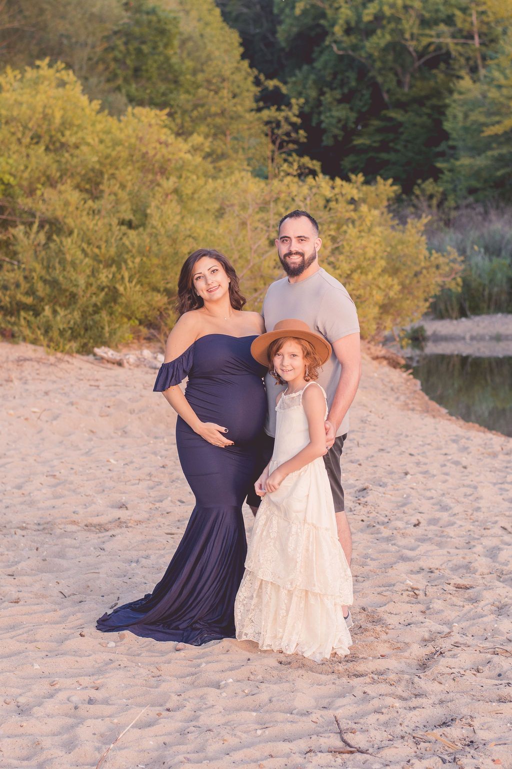 Pregnant woman, man, and young girl posing on a sandy beach at sunset with trees behind them