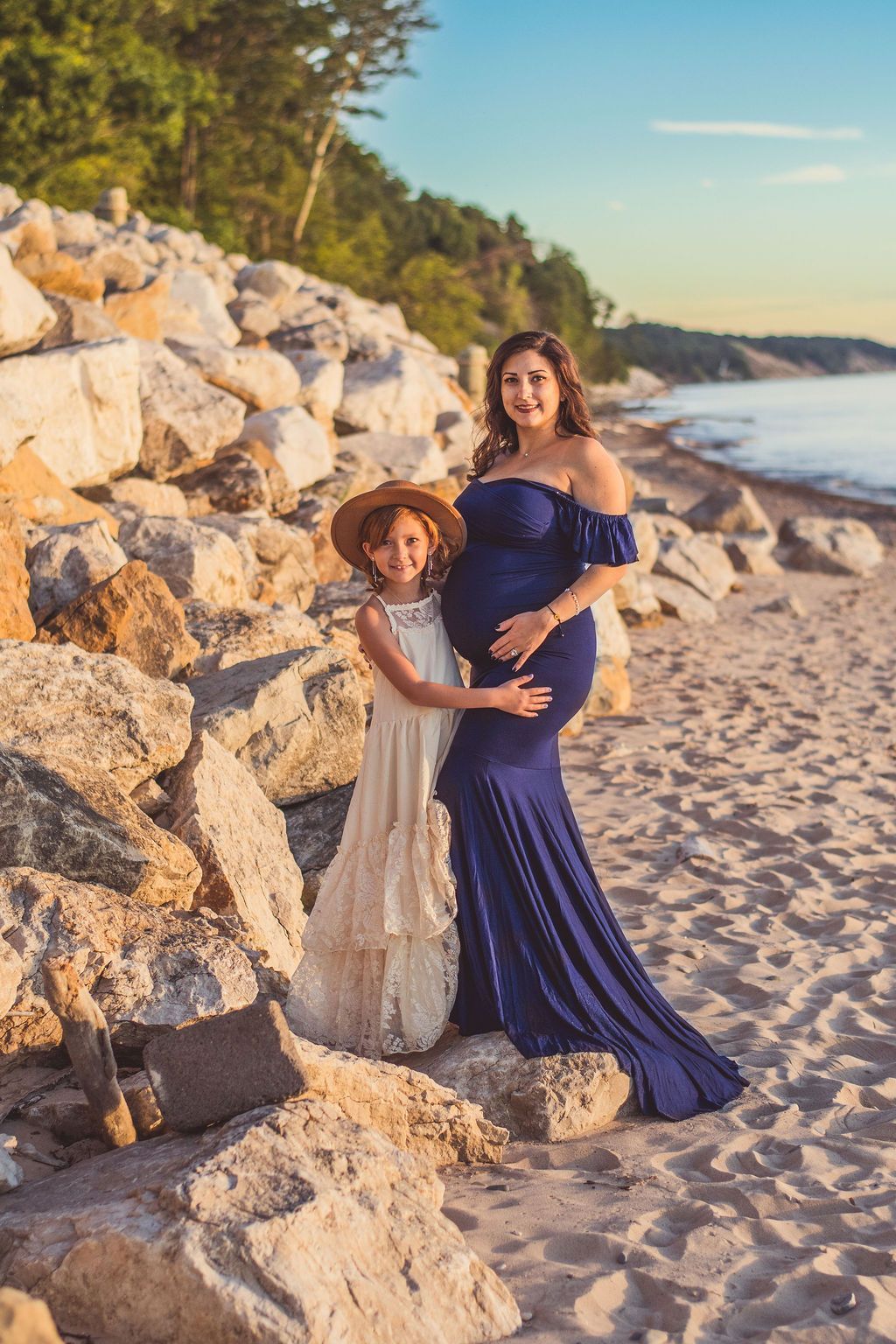 Two women in blue and white dresses posing on a sandy beach beside large rocks at sunset