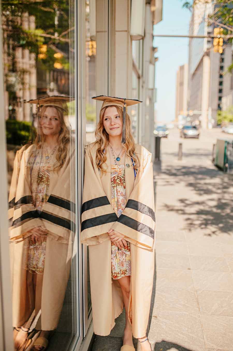 Woman in a striped coat and dress standing by a mirrored storefront on a sunny sidewalk