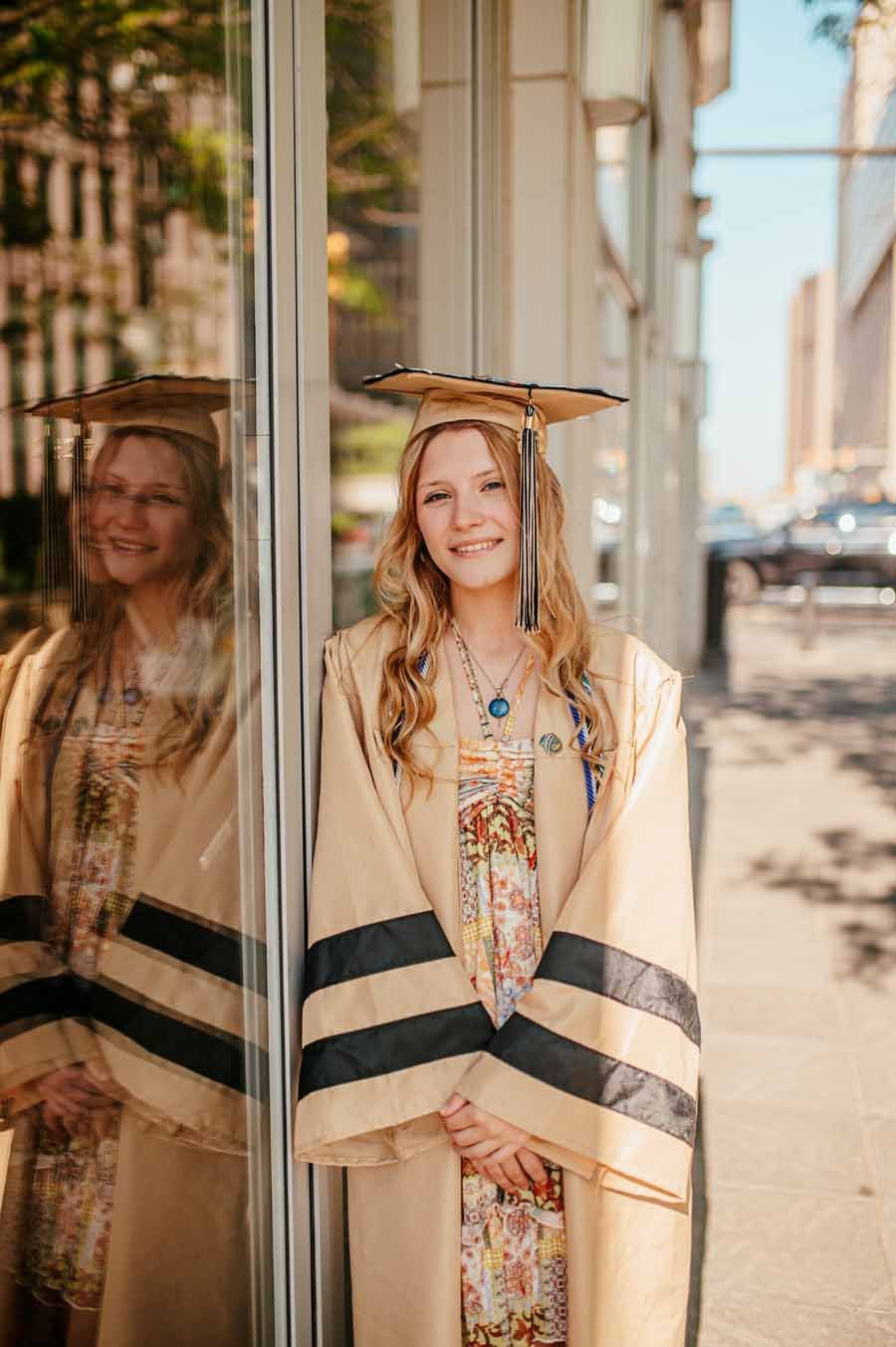 Graduate in cap and gown standing by a reflective wall outdoors, smiling in sunlight