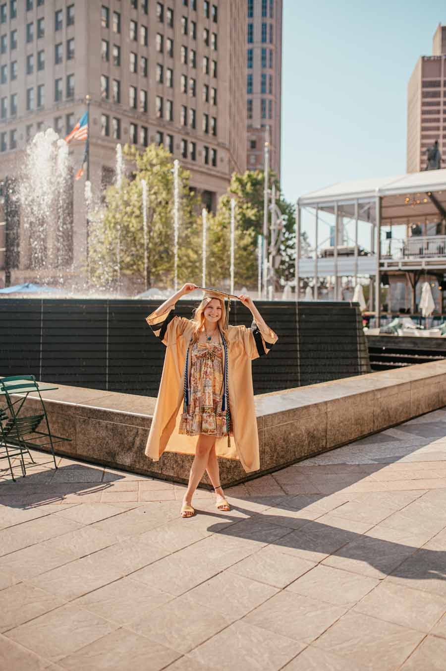 Person in a tan coat walking near a fountain in a city plaza, with buildings in the background.
