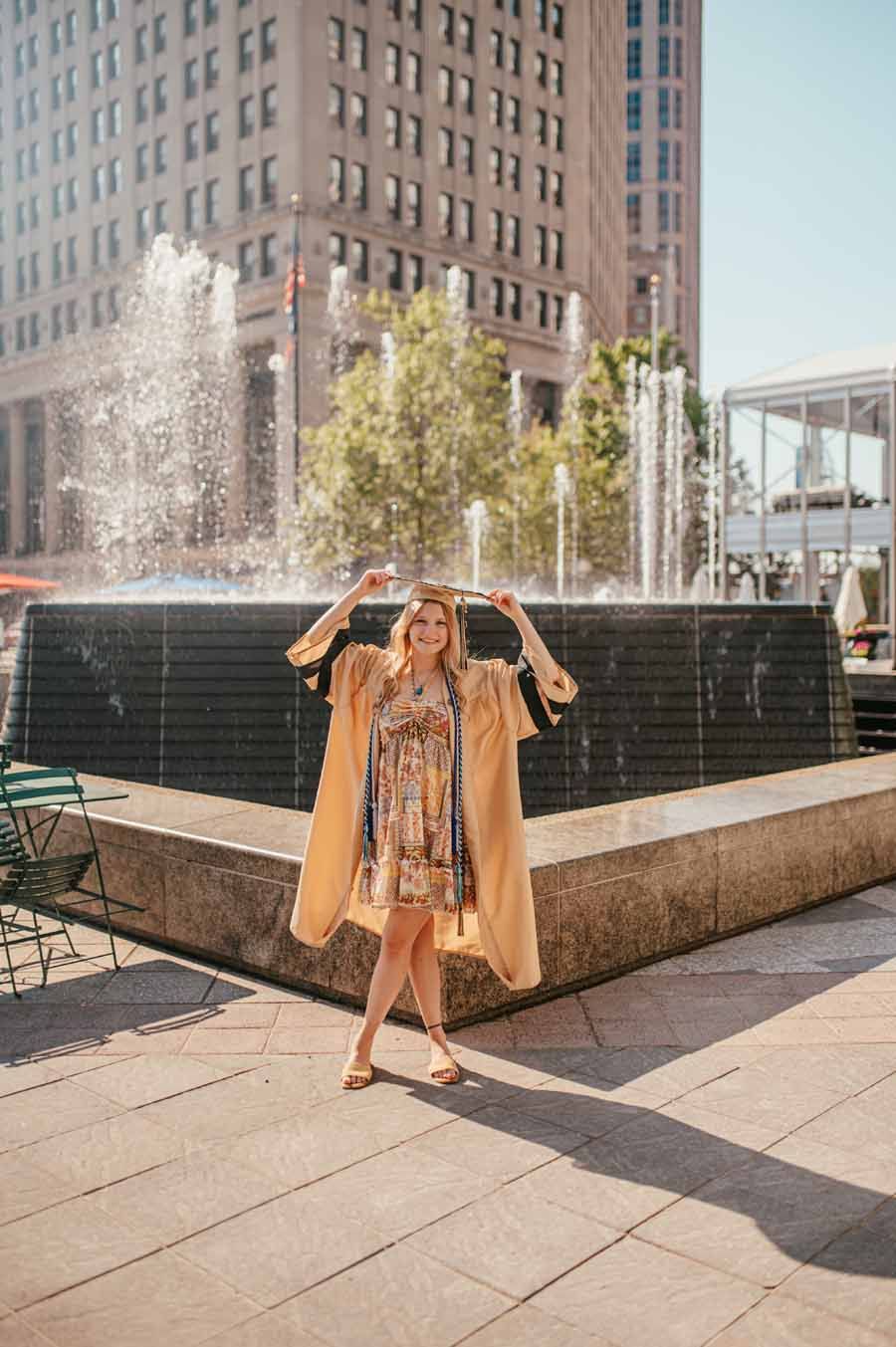 Woman posing by a fountain in a city plaza, wearing a tan coat and patterned dress.