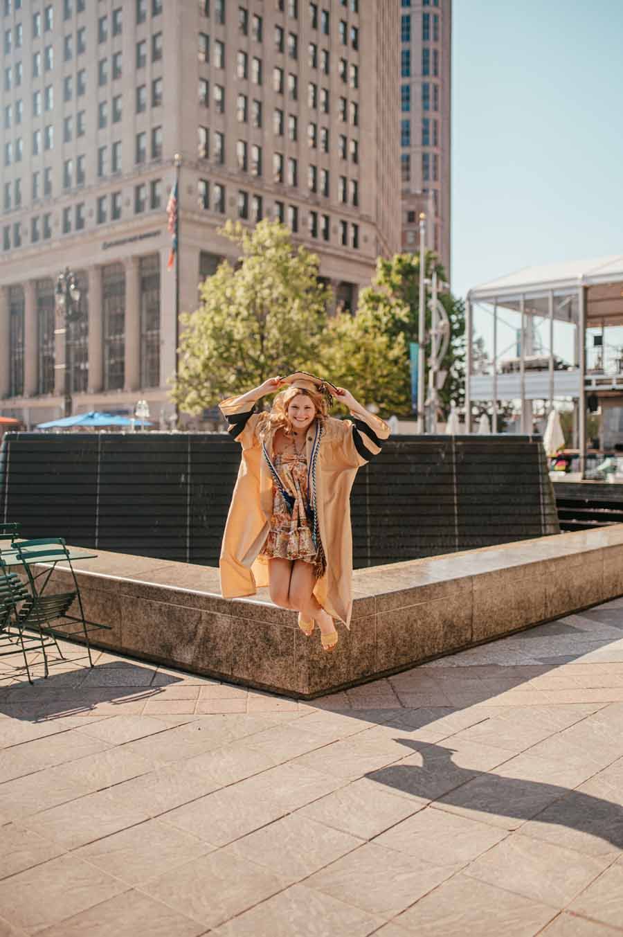 Woman in beige coat posing by a planter in a city plaza with tall buildings behind her