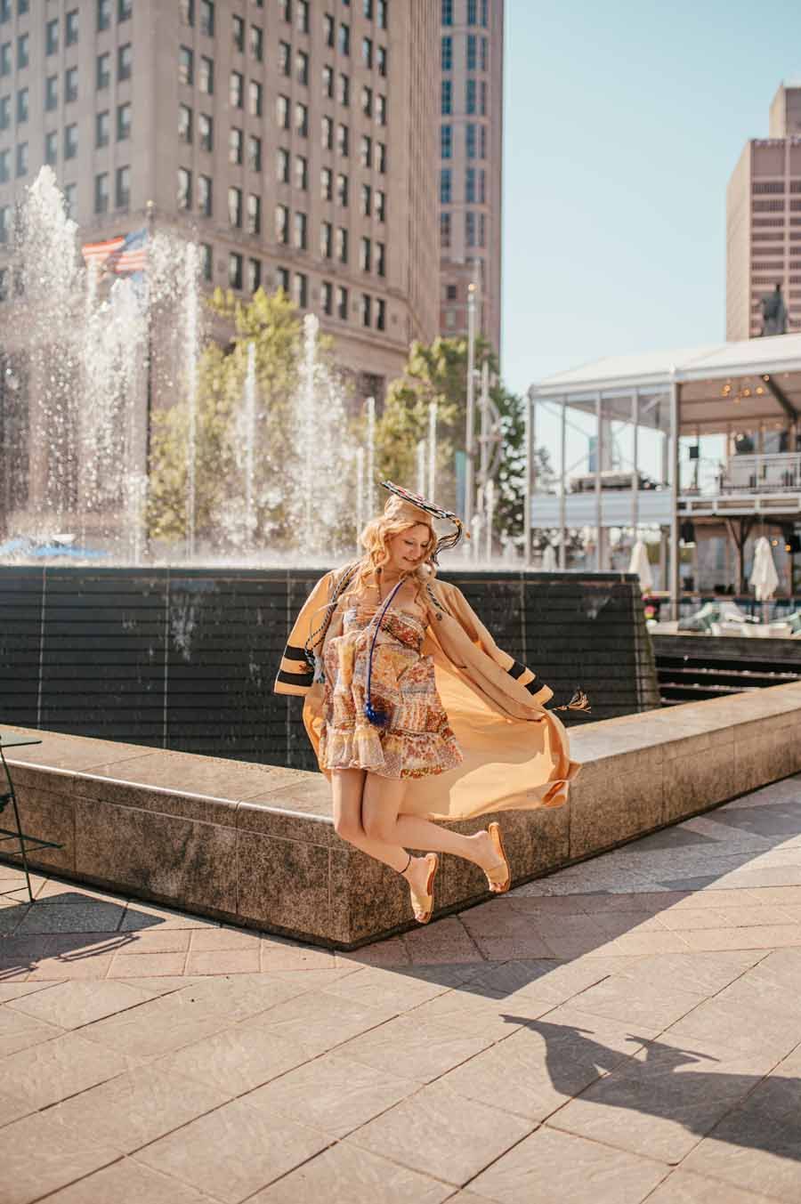 Person in a tan coat posing by a city fountain on a sunny day