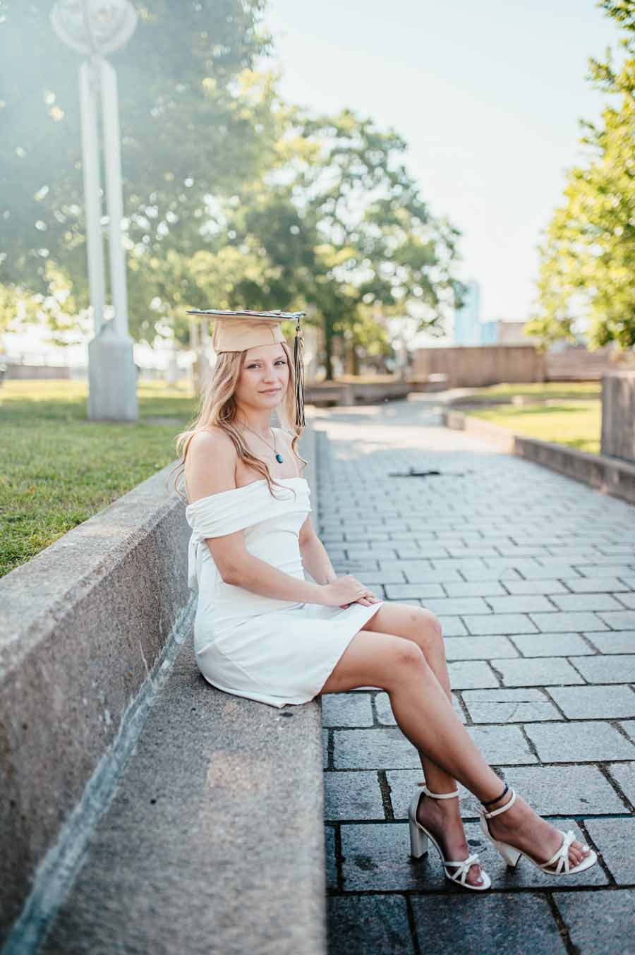 Woman in a white dress sitting on a stone wall along a paved walkway in sunlight.