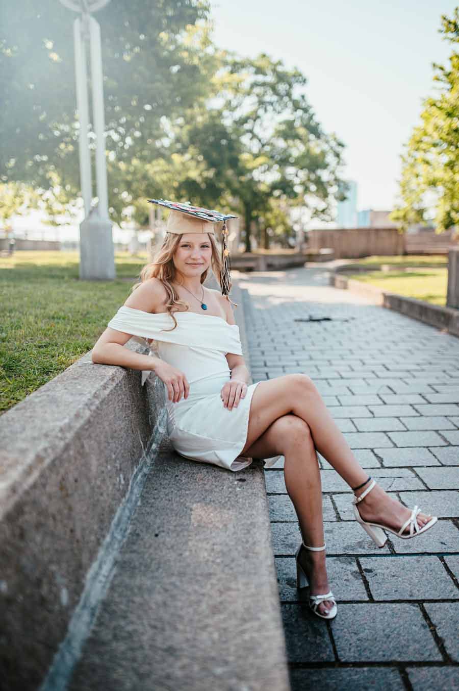 Woman in a white dress sits on a low wall on a sunlit paved walkway, smiling.
