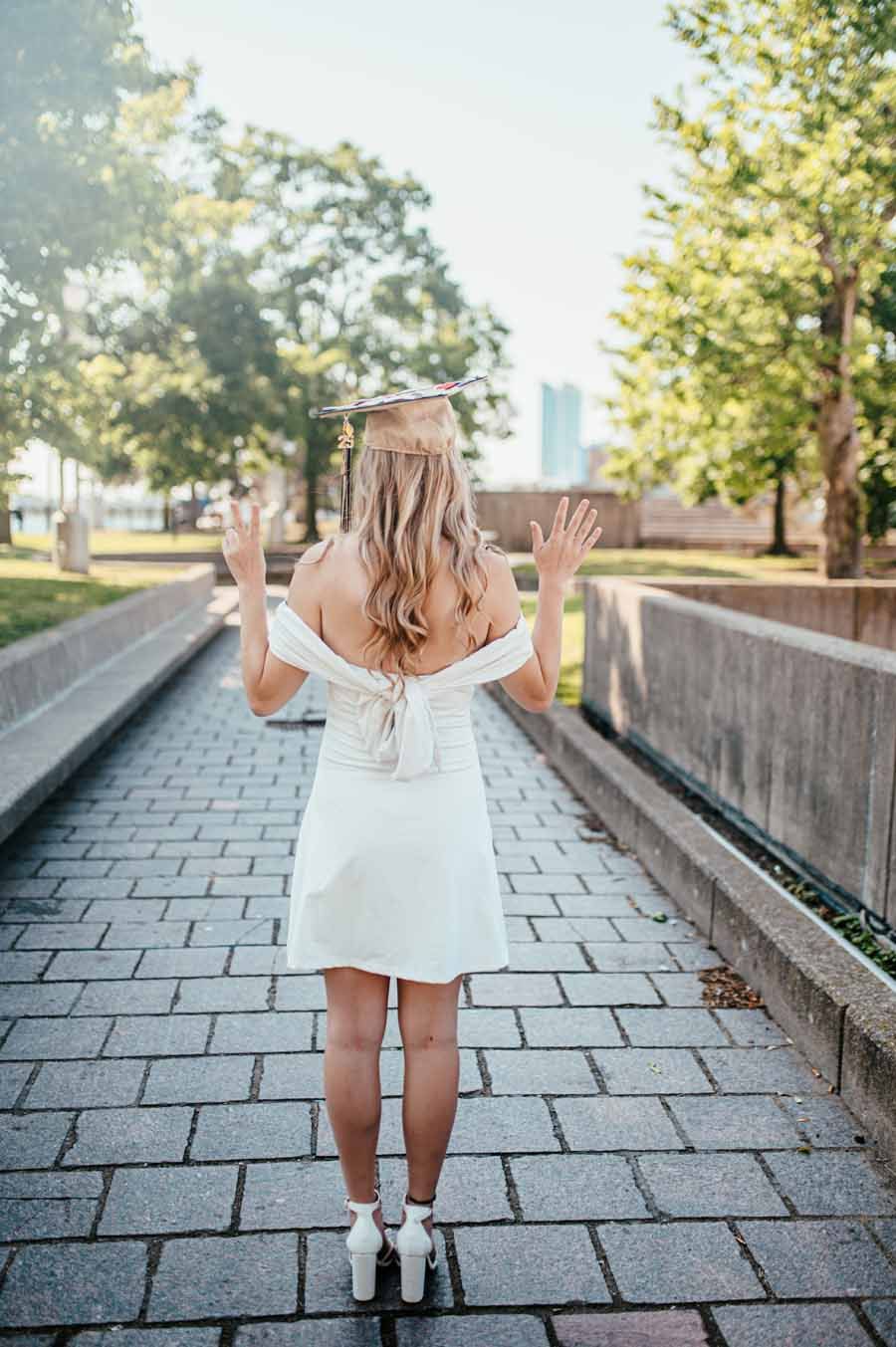 Person in a white dress standing on a stone path with hands raised, framed by trees in sunlight.
