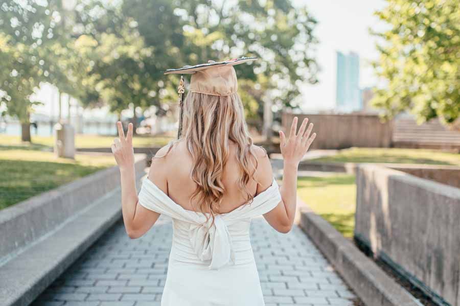 Person in a white dress with arms raised, walking down a tree-lined path outdoors.