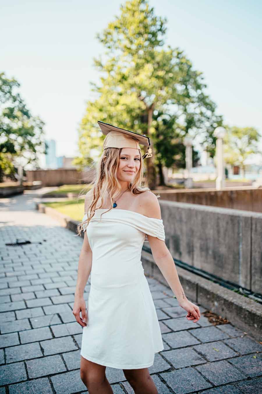 Woman in a white dress walking outdoors on a paved path near a wall and trees, smiling in sunlight