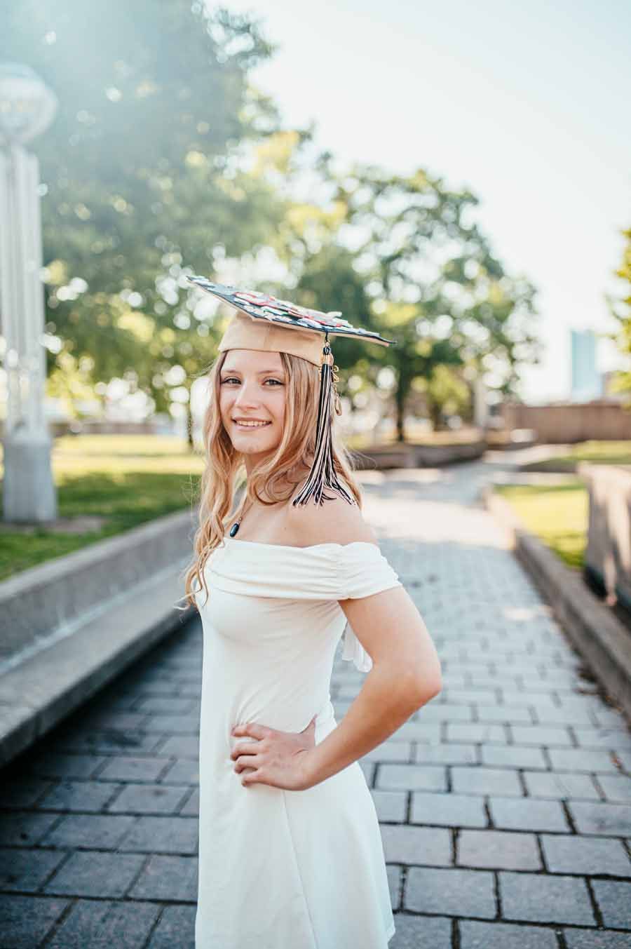 Smiling woman in a white dress and graduation cap standing outdoors on a brick path