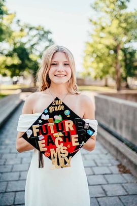 Person in white dress holding a black graduation cap with gold letters, standing outdoors and smiling