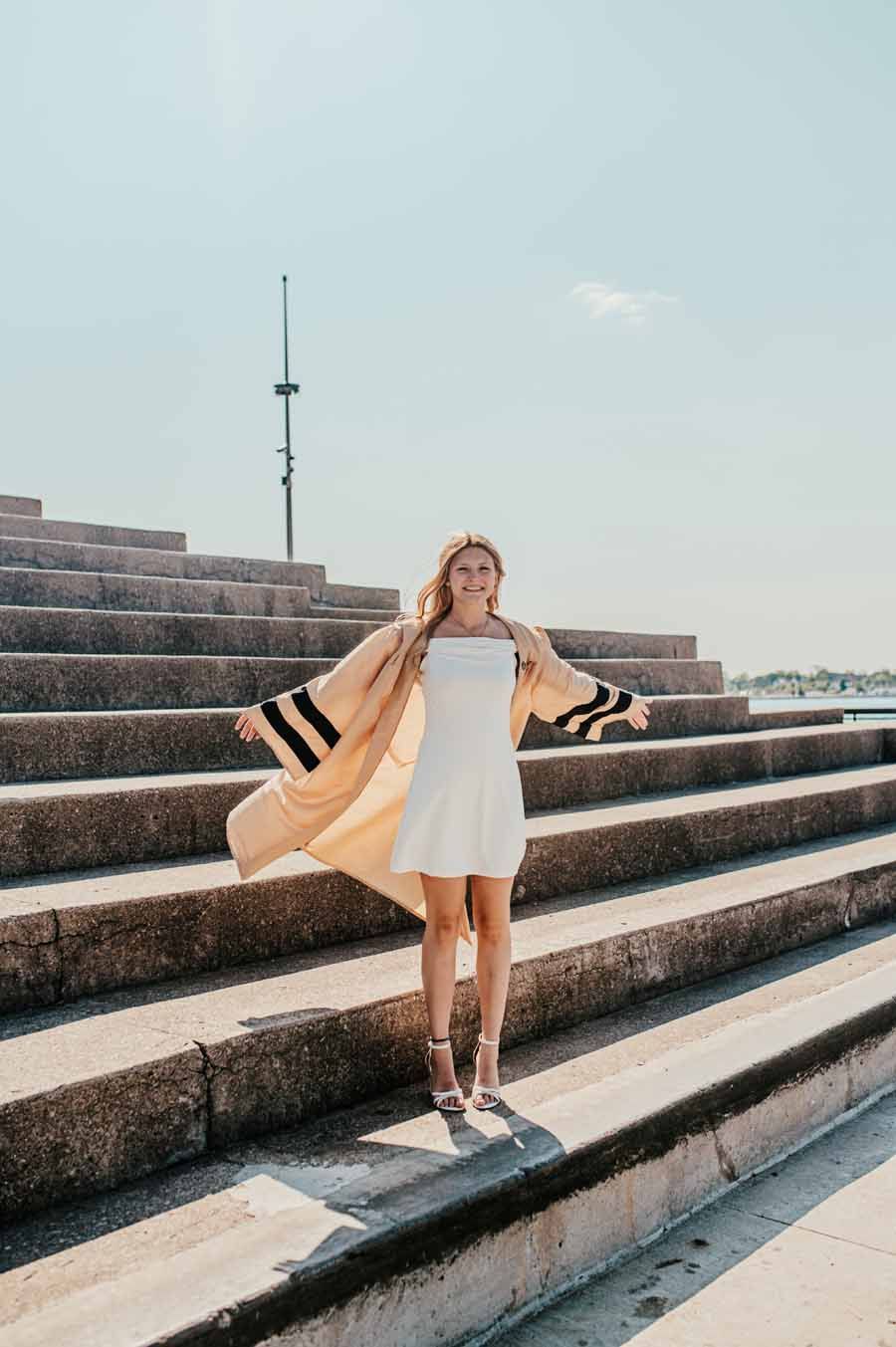 Woman in a white dress and tan coat posing on outdoor concrete steps under a clear sky