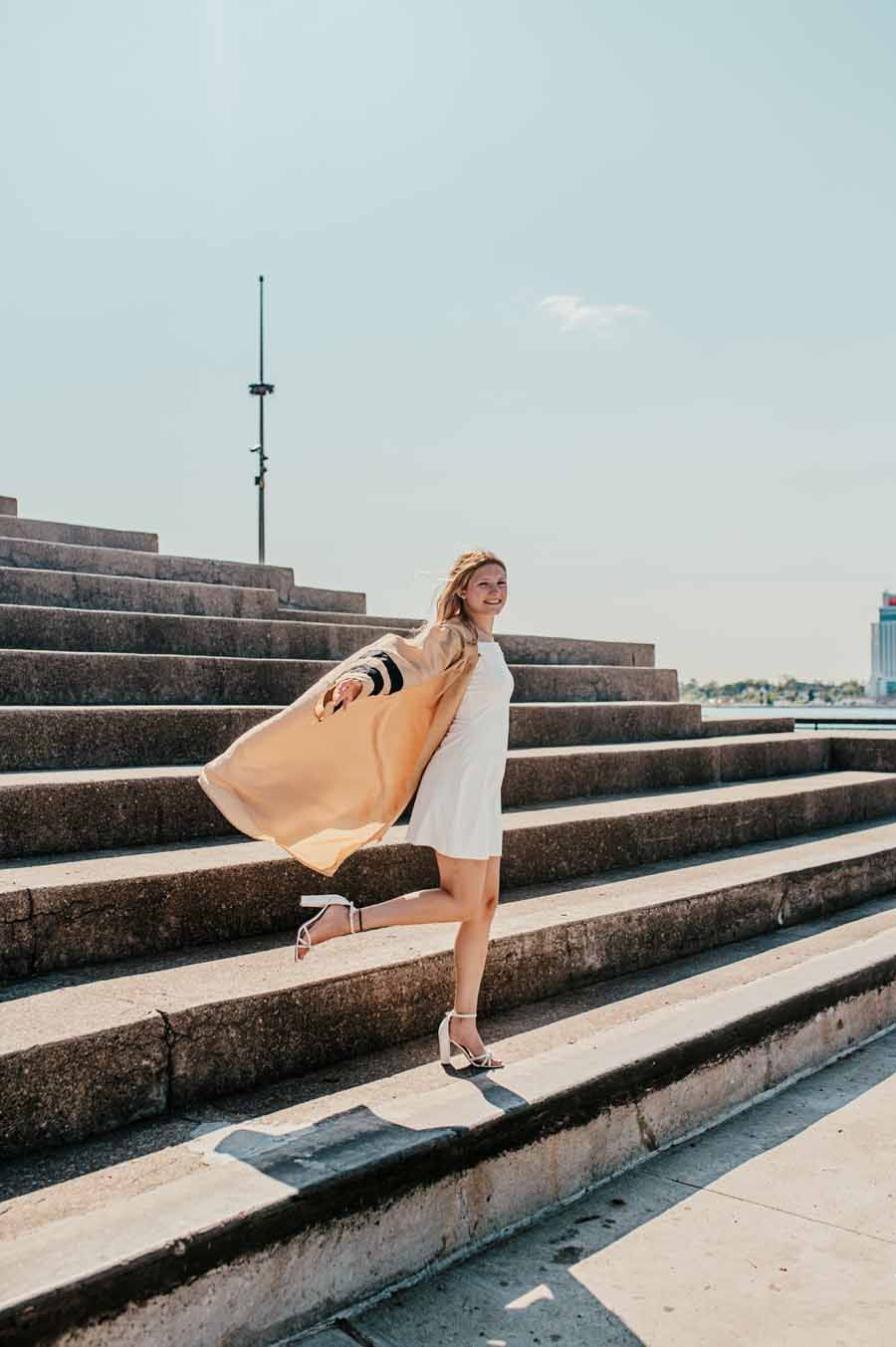 Woman in a white dress and tan coat posing on stepped seating outdoors under a clear sky