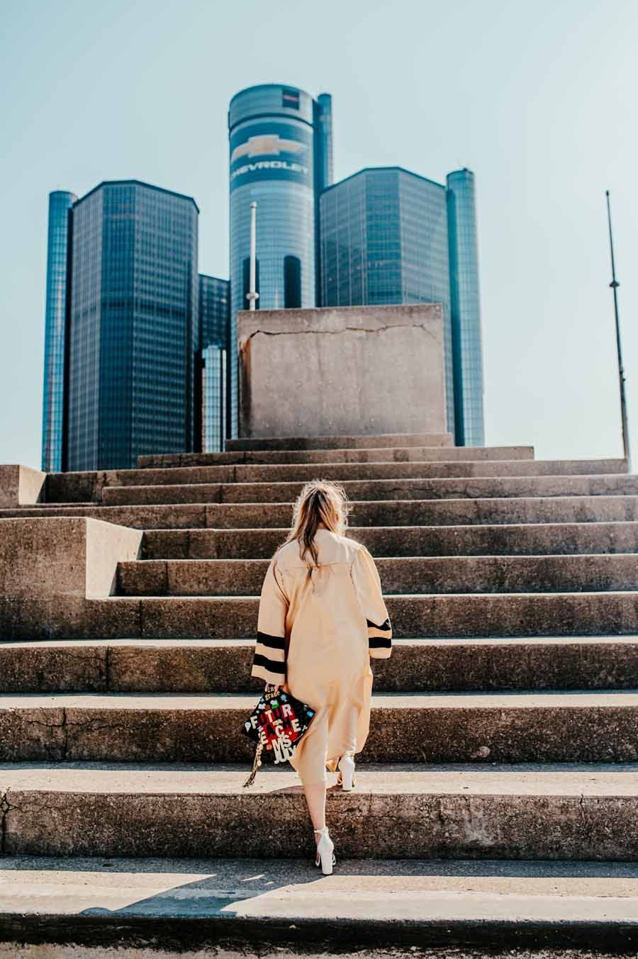 Person in a beige coat climbing concrete steps toward modern glass skyscrapers.