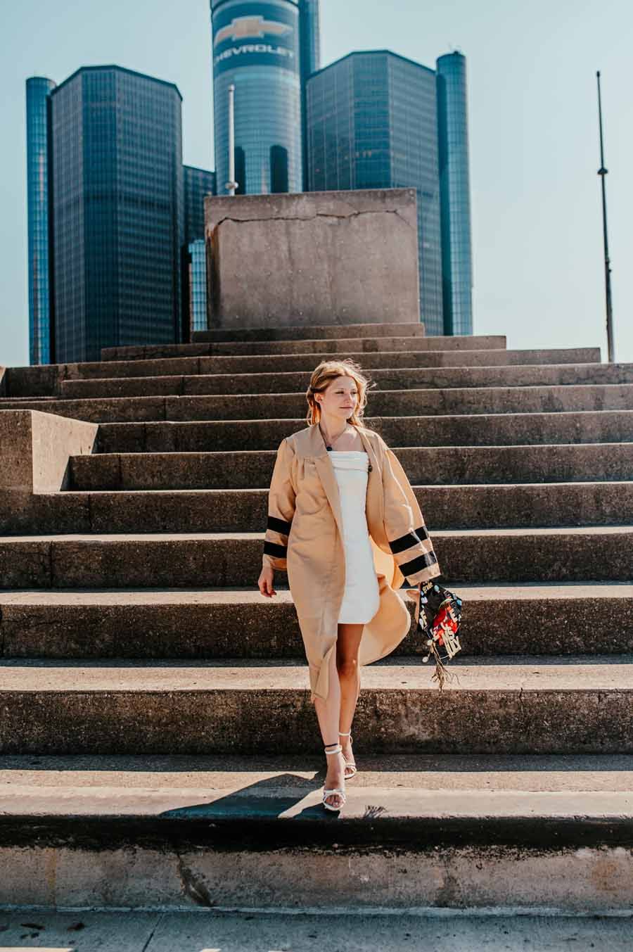 Person standing on wide steps in front of modern skyscrapers, wearing a tan coat and holding flowers.